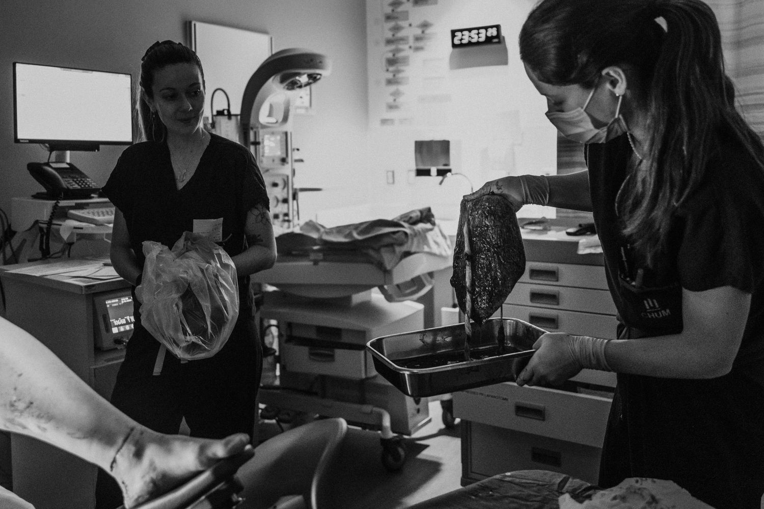 Healthcare workers in dark scrubs attend to a patient in a medical treatment room, captured in black and white photography.