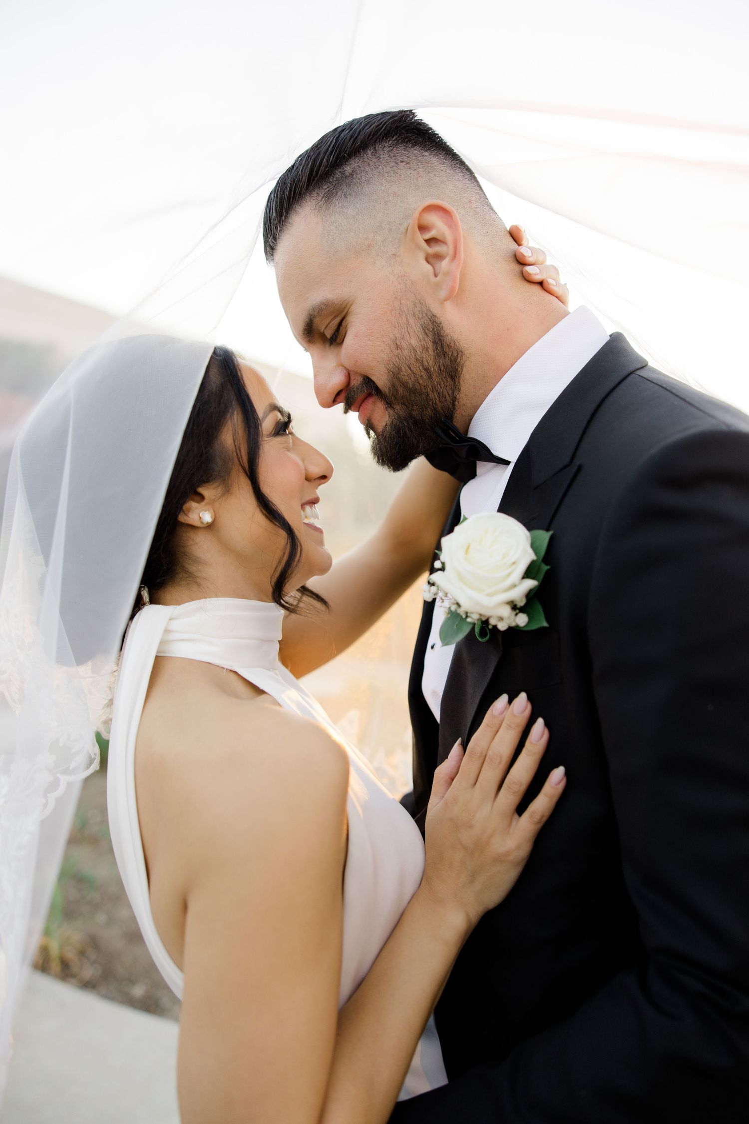 A bride and groom share a romantic moment under a wedding veil, smiling at each other in formal attire.