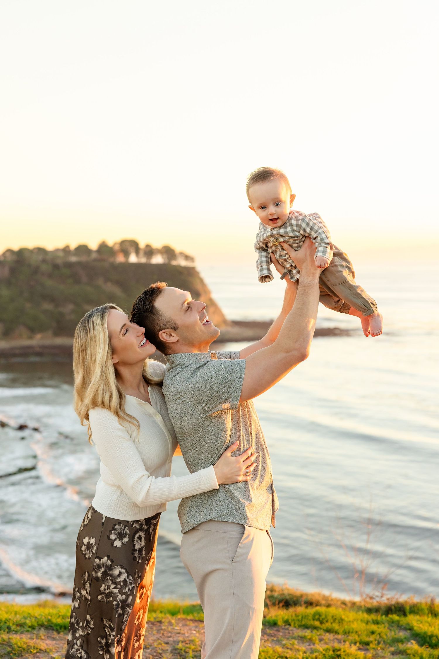 A happy family poses at sunset by the ocean, with a man lifting a smiling baby while a woman stands beside them.