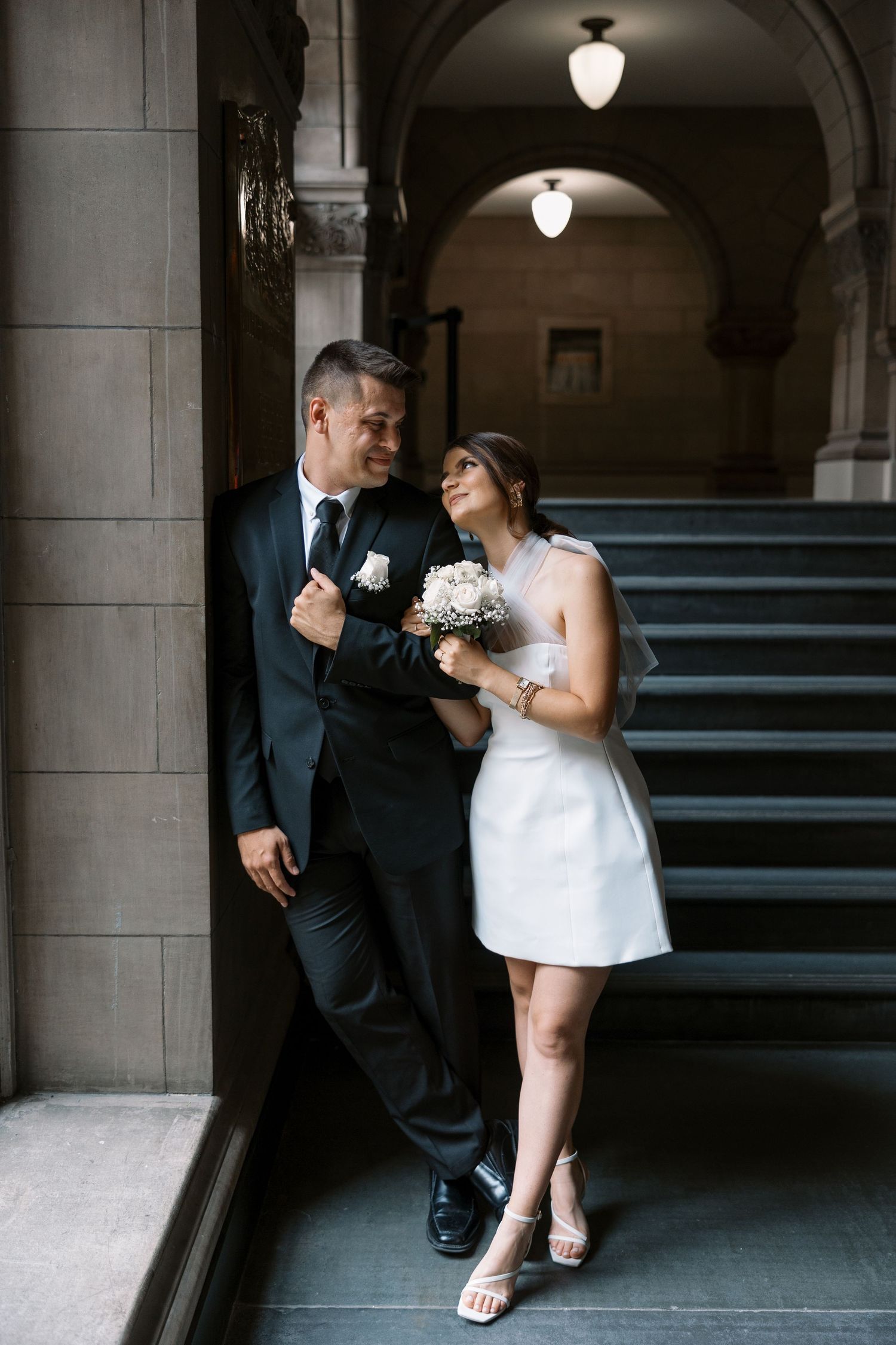 A couple embraces in formal attire near an ornate stone staircase with architectural columns.