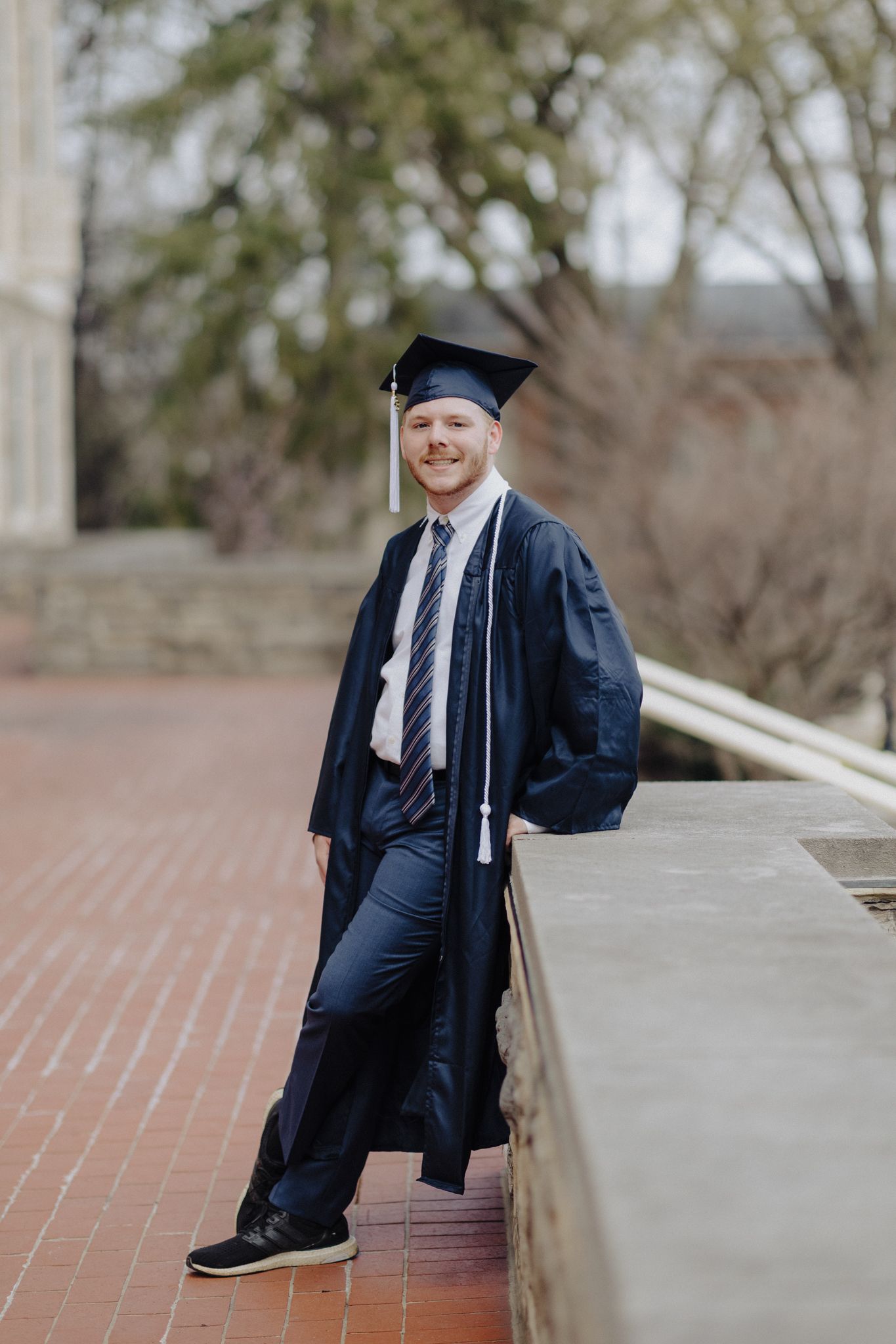 Graduate in traditional navy regalia poses on brick pathway with bright smile.