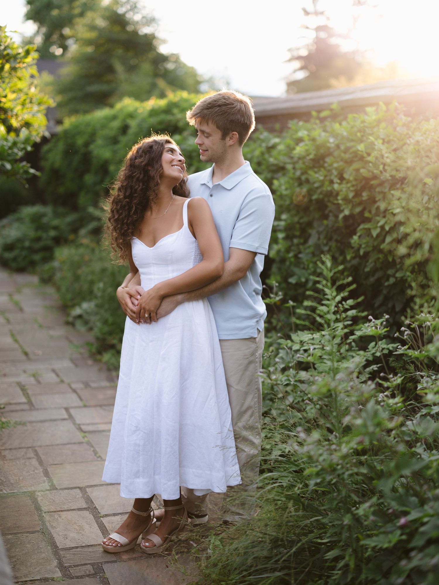 Couple embracing on a garden path at golden hour, woman in white dress, man in light blue shirt, lush greenery background.