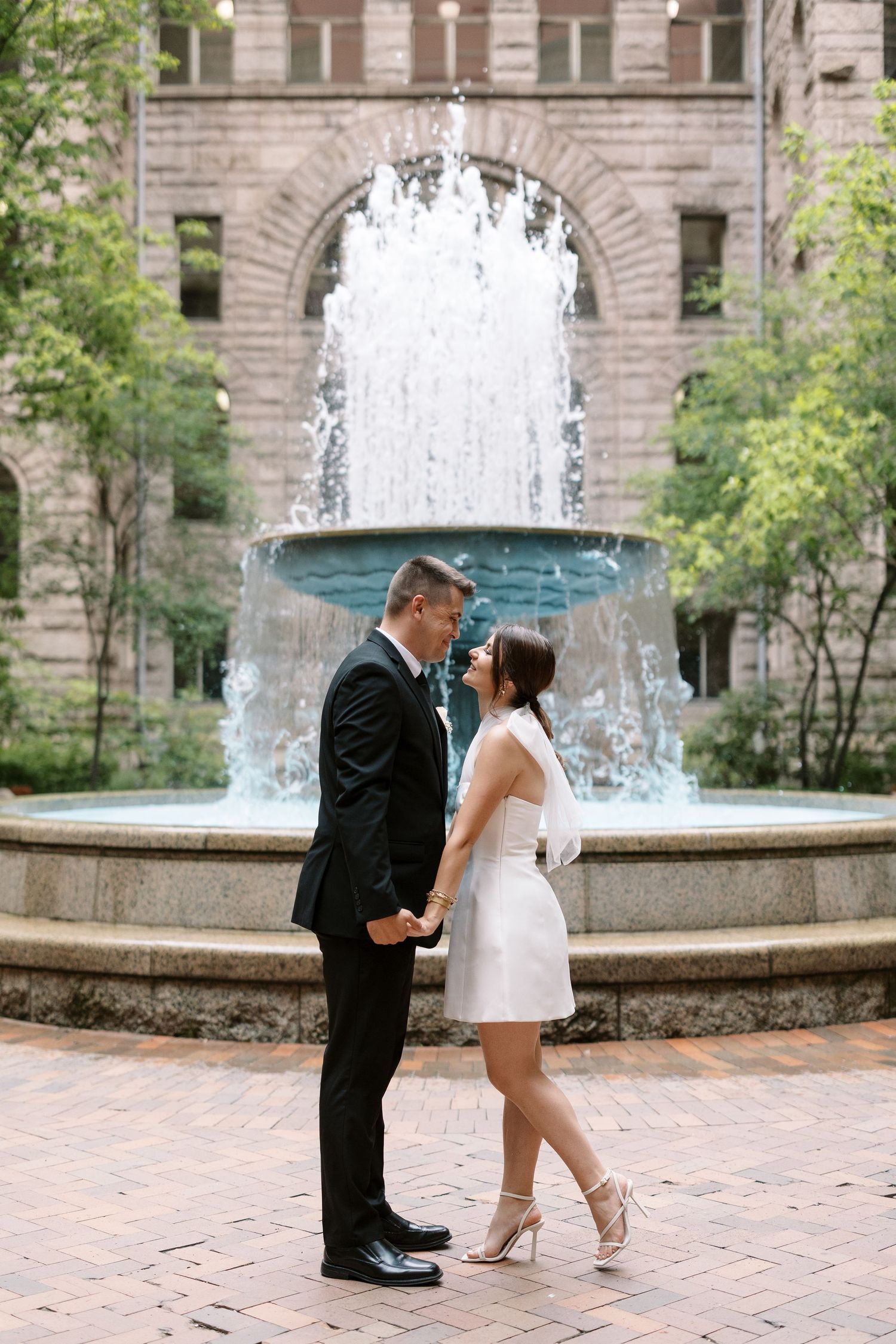A couple shares an intimate moment in front of a beautiful flowing fountain on a brick courtyard.
