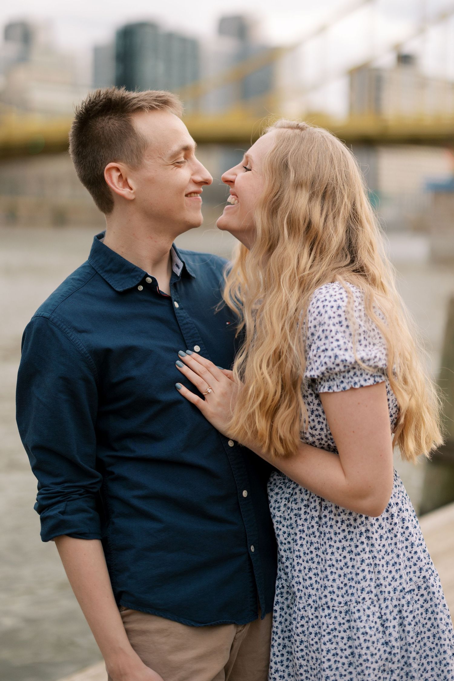 A candid moment of a couple laughing together with a bridge and cityscape in the background.