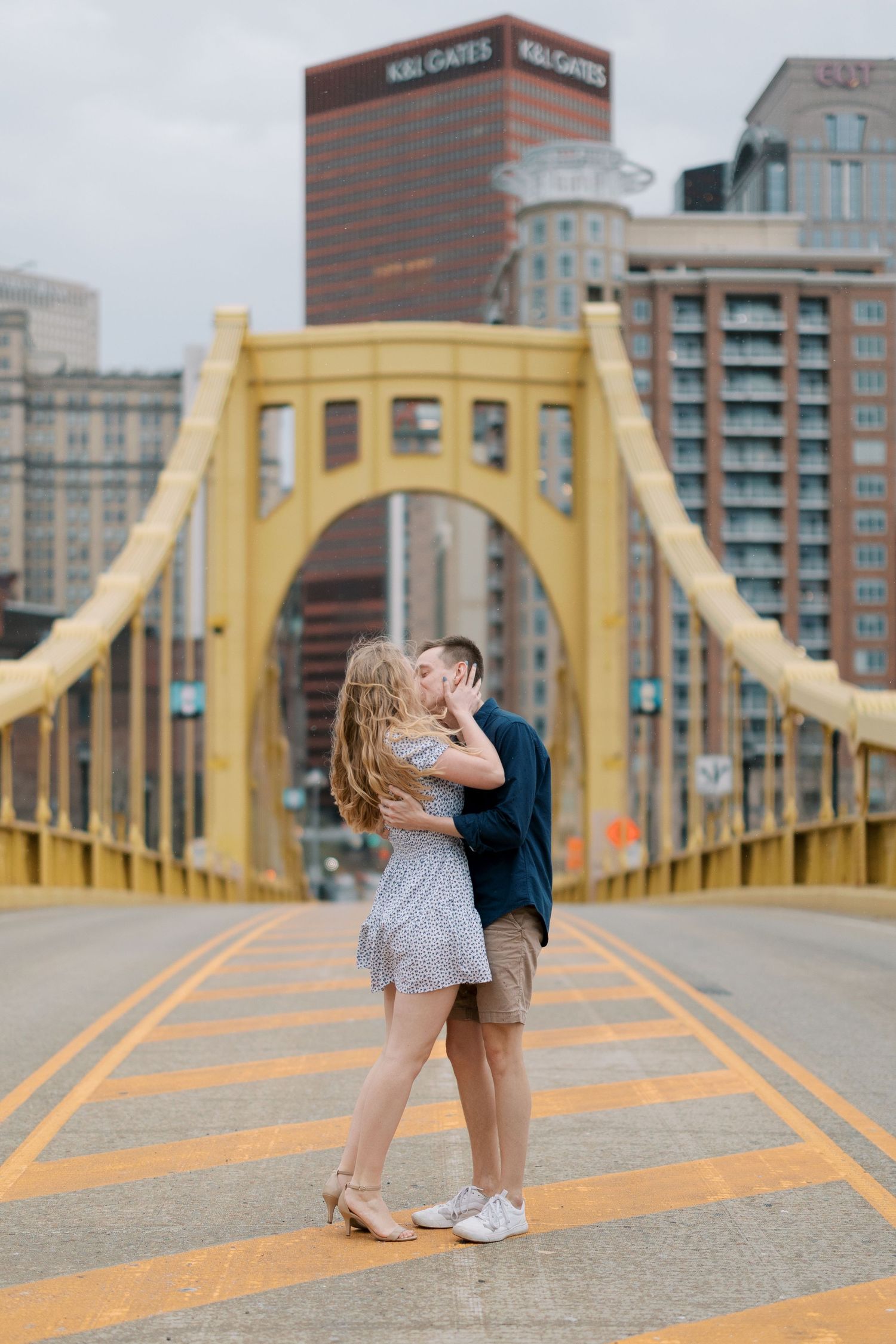 Couple embracing on iconic yellow bridge with Pittsburgh skyline in background.