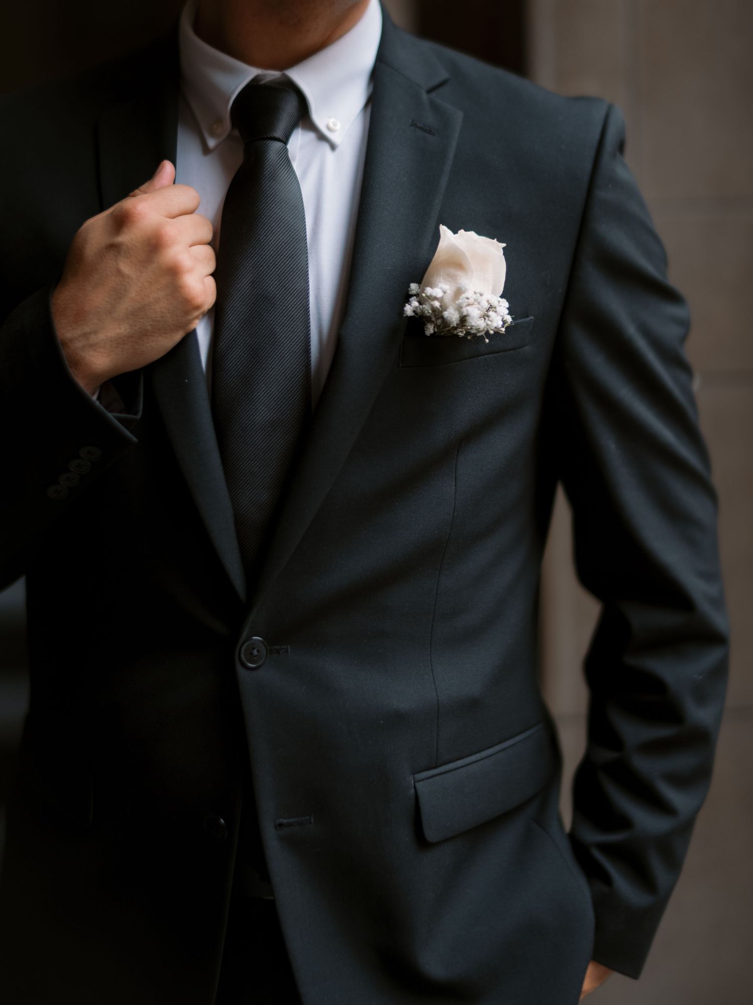 A detailed shot of a dark suit jacket with white boutonniere and hand adjusting the tie.