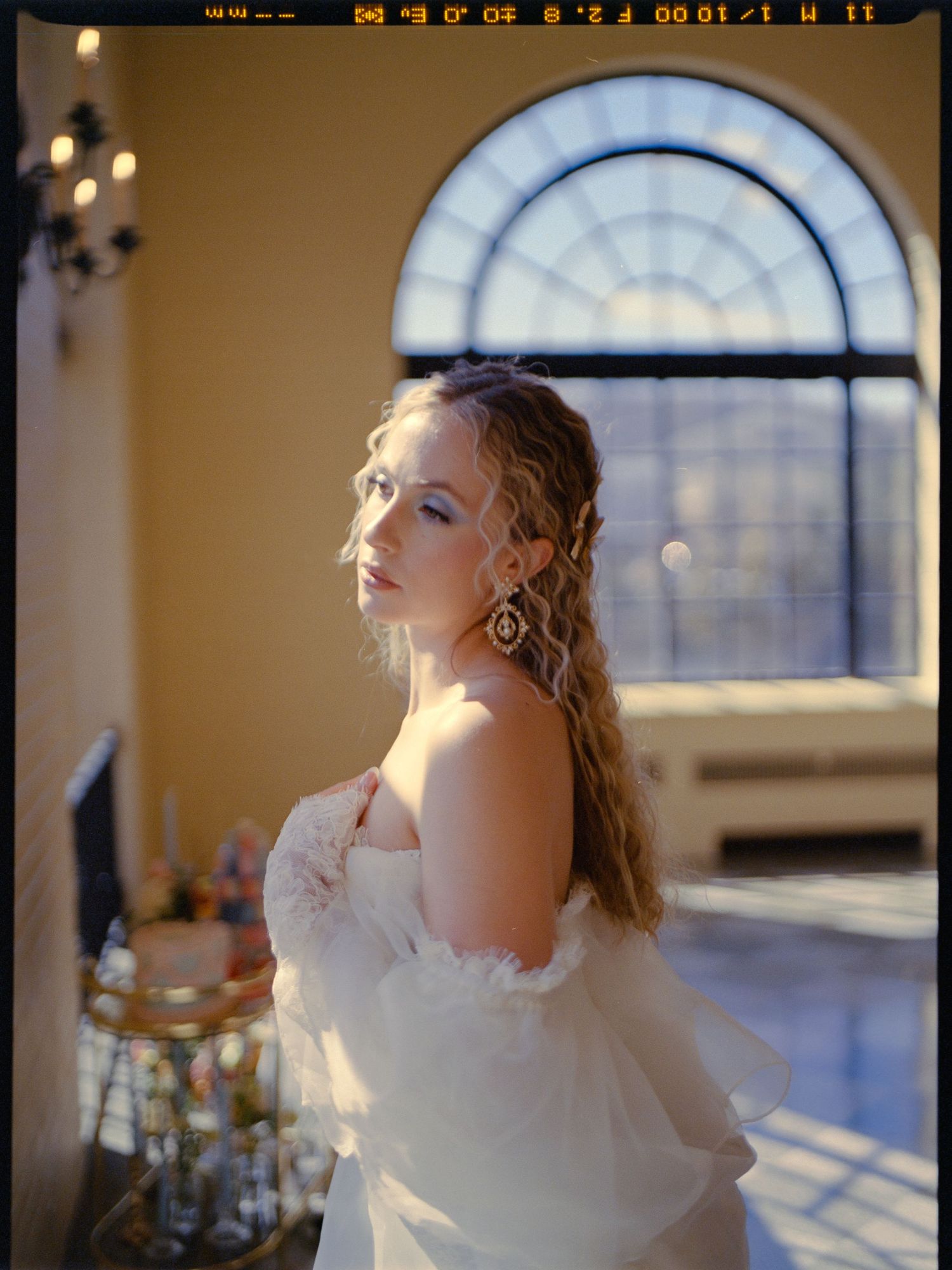 A bride in an elegant off-shoulder gown poses near an arched window with natural light.