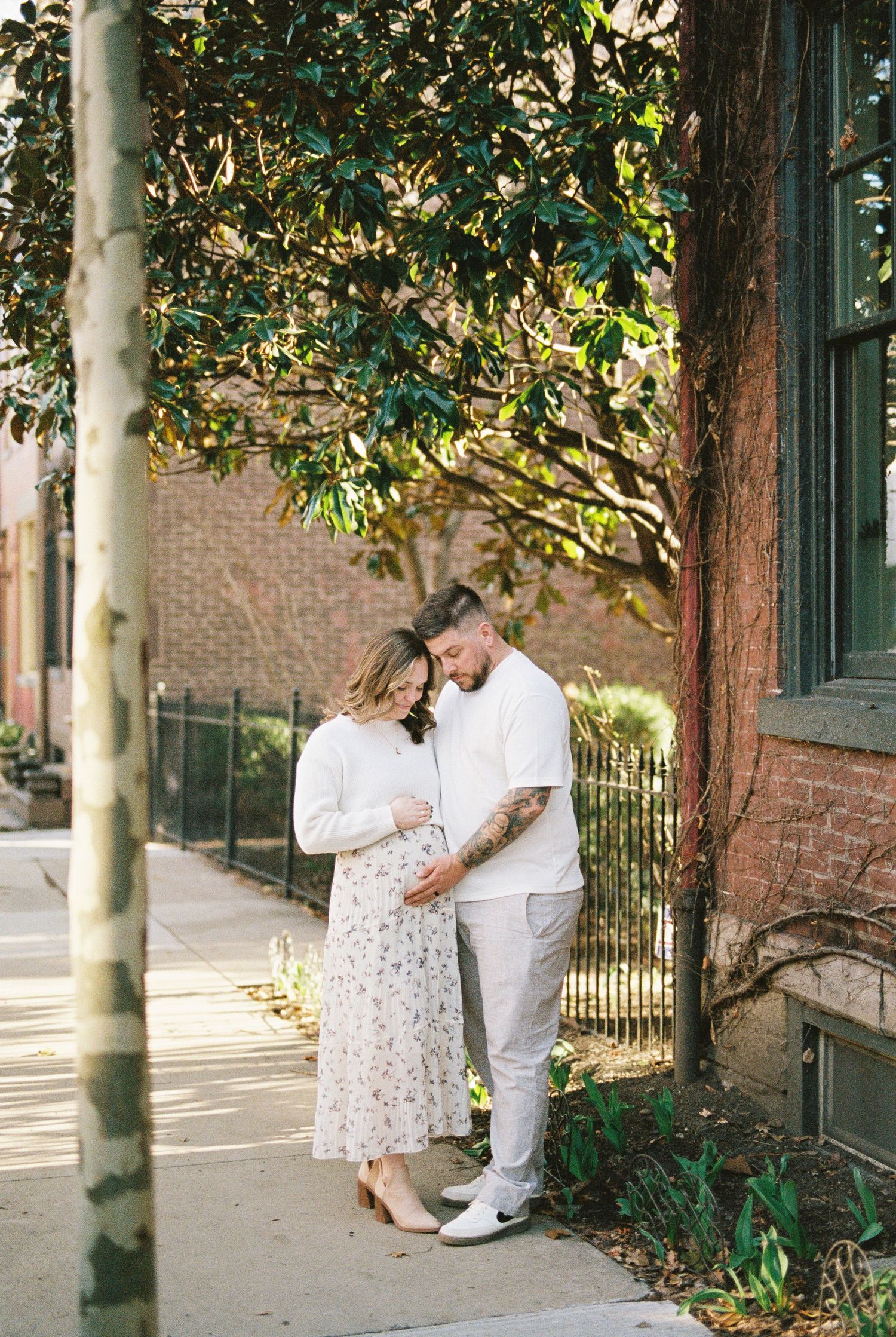 A romantic couple in white outfits share loving moments against a brick wall with trees in an urban setting.