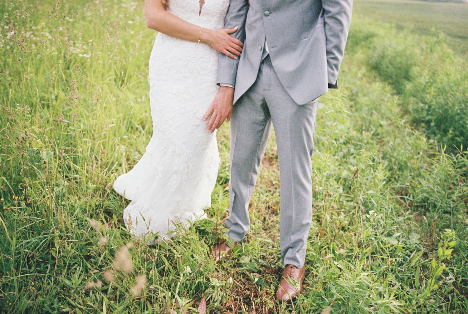 A couple in wedding attire walk through a green meadow with sunlight filtering through the grass.