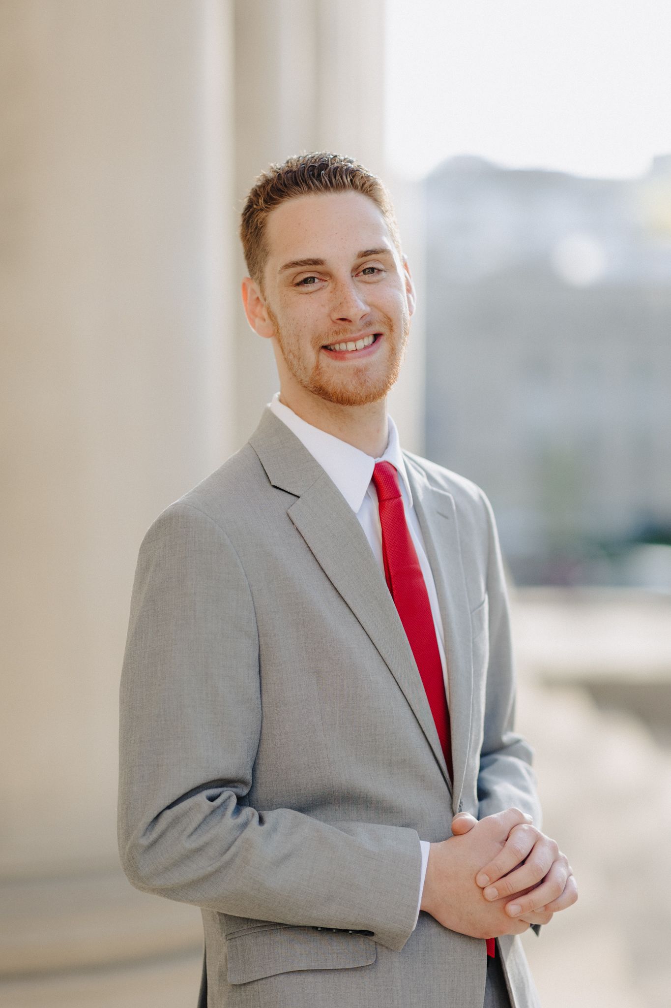 Professional headshot of person in grey suit and red tie against architectural background.
