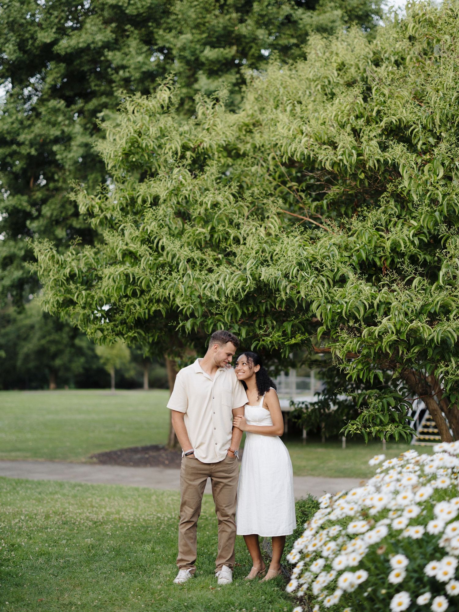 A couple poses together outdoors in a park setting with flowering plants and green trees in the background.