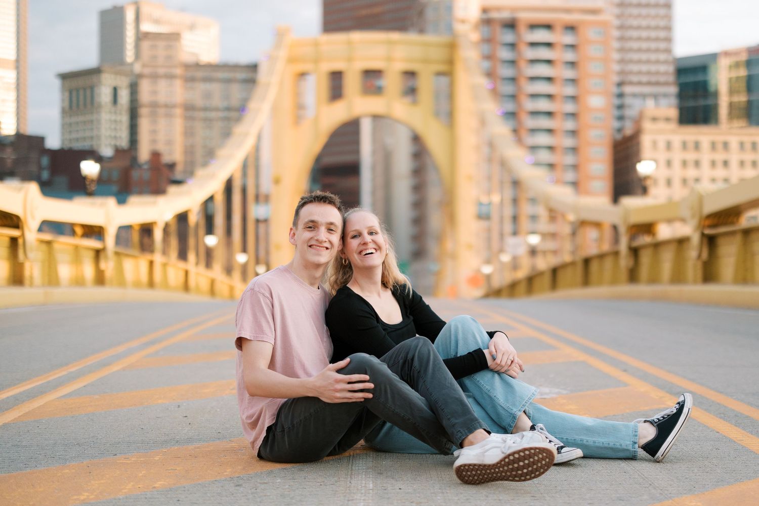 A couple sitting casually on a yellow bridge with Pittsburgh's skyline in the background.
