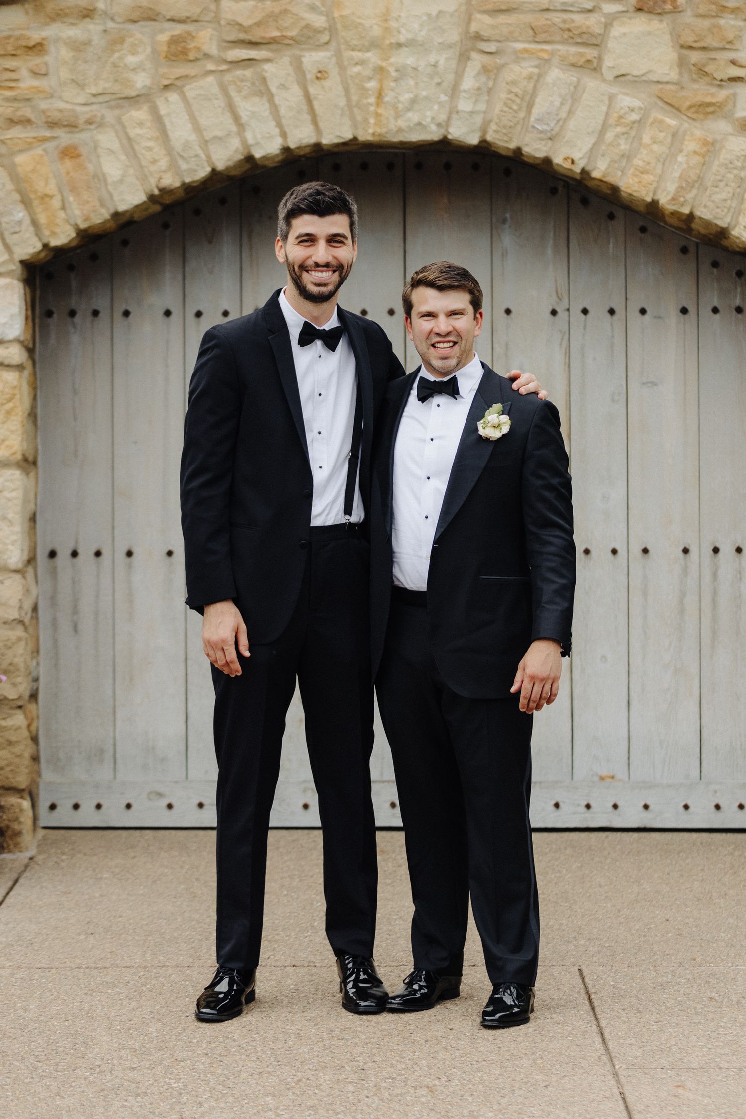 Two people in black tuxedos stand together smiling in front of a stone arch doorway.