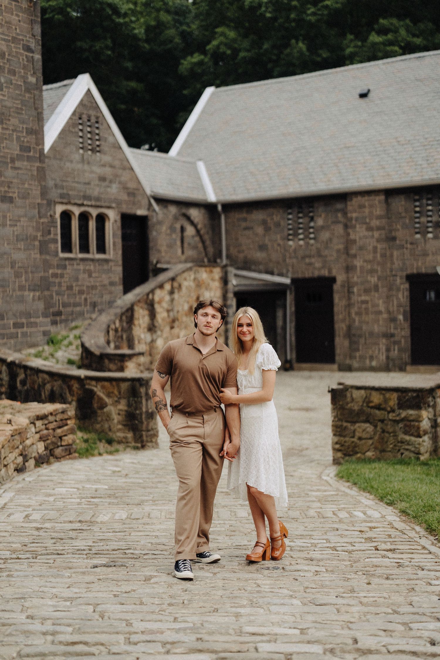 A couple poses together on a cobblestone path in front of a rustic stone building.