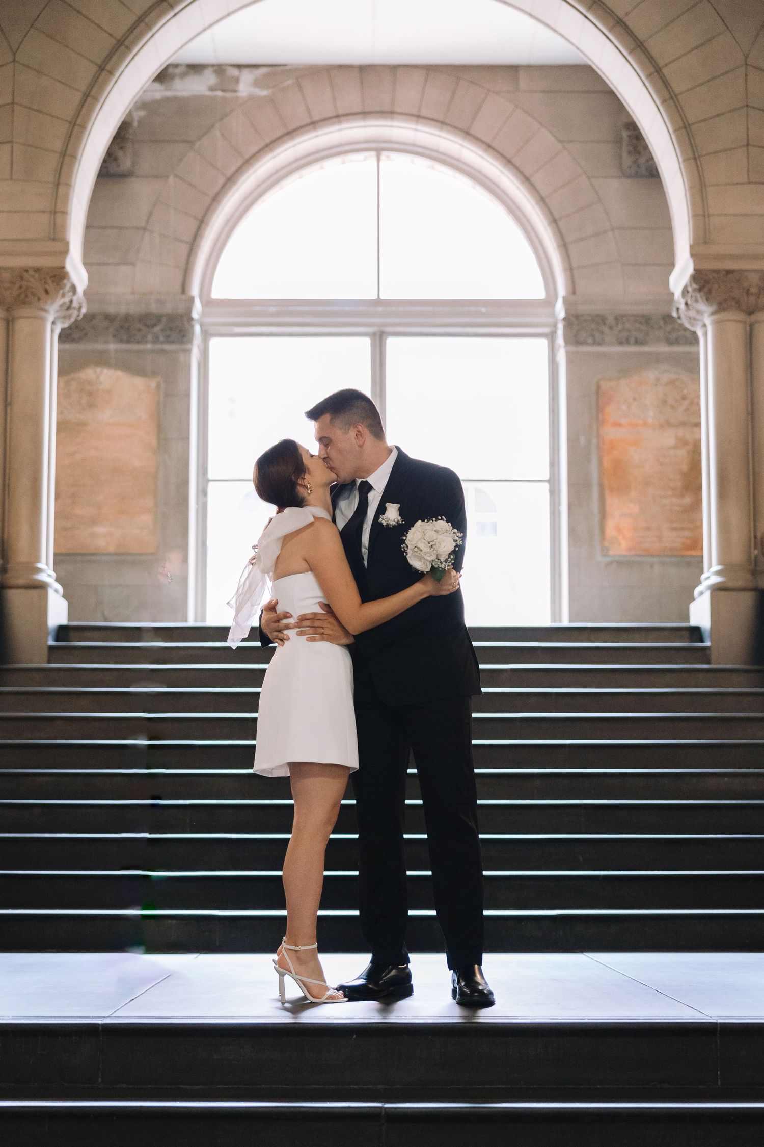 A couple embraces on grand stone steps beneath an ornate archway in formal attire.