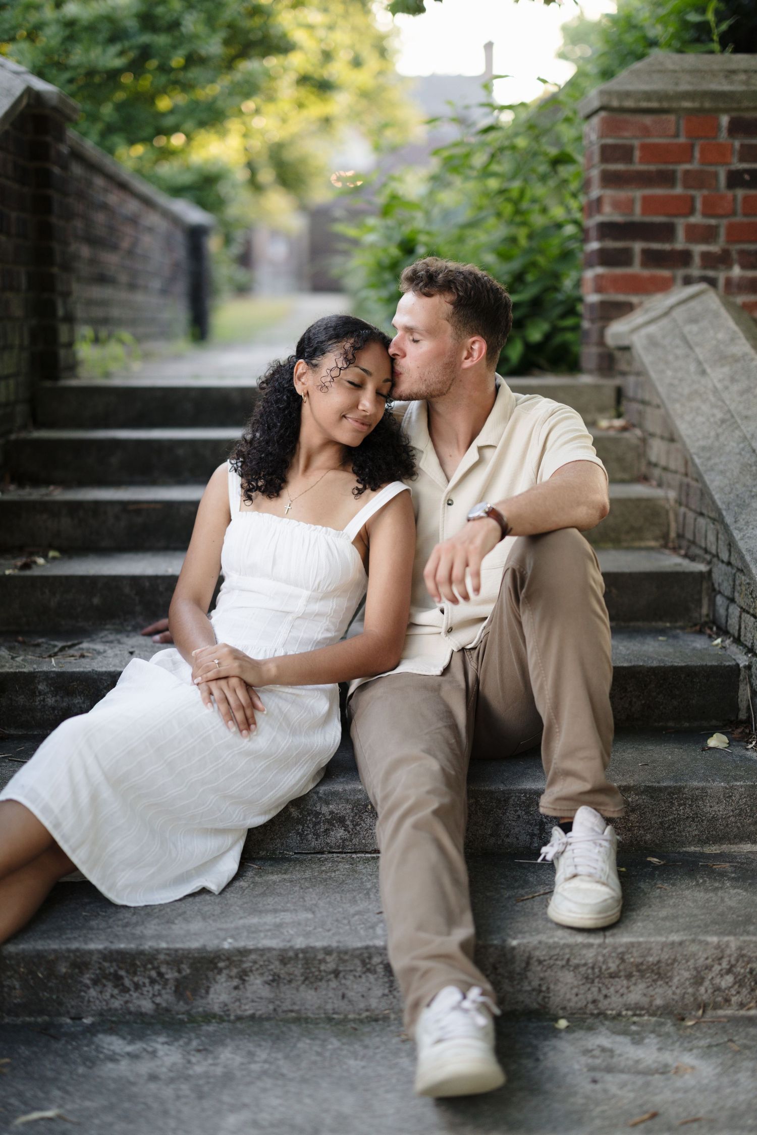 A romantic couple shares tender moments while sitting on stone steps amidst a garden setting.