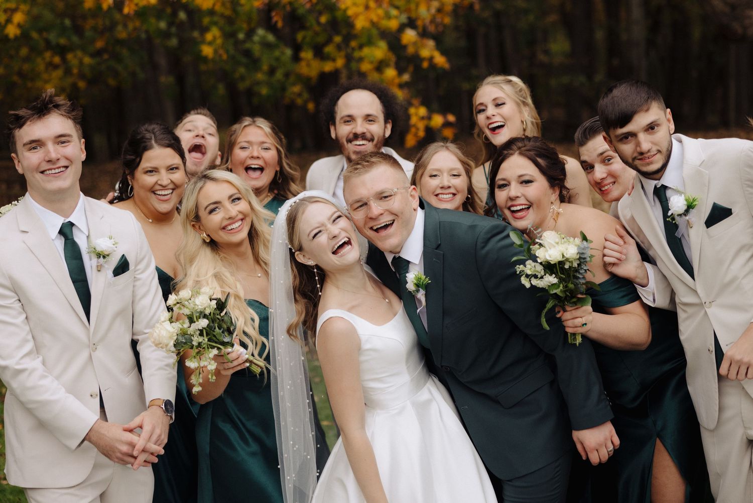 Wedding party poses together outdoors with autumn foliage in background.