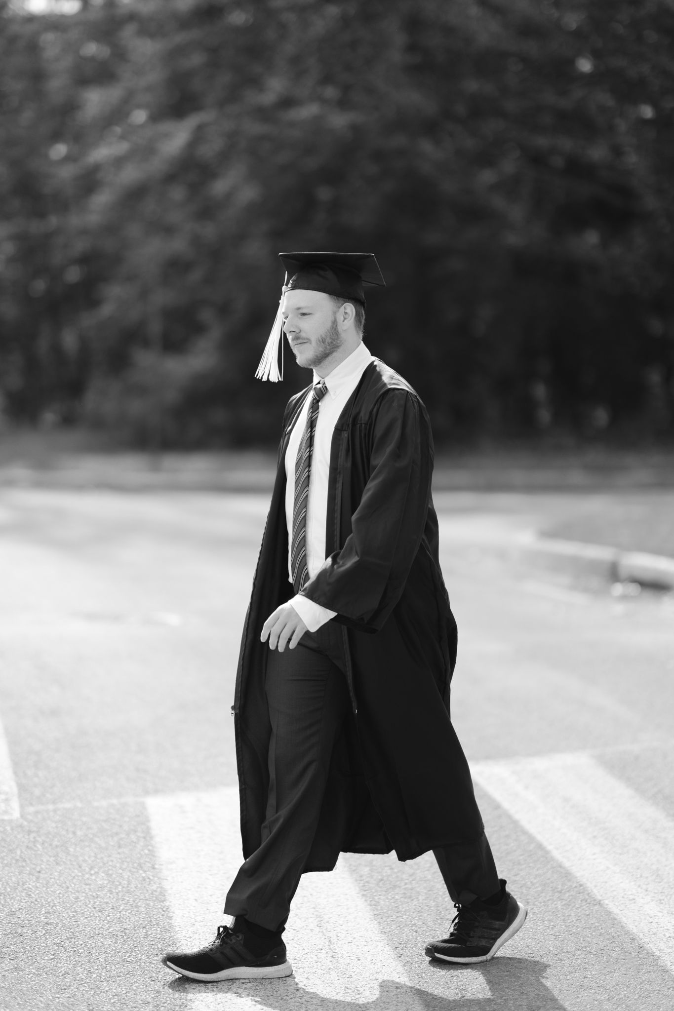 Graduate in black cap and gown walks confidently across a path in black and white photography.