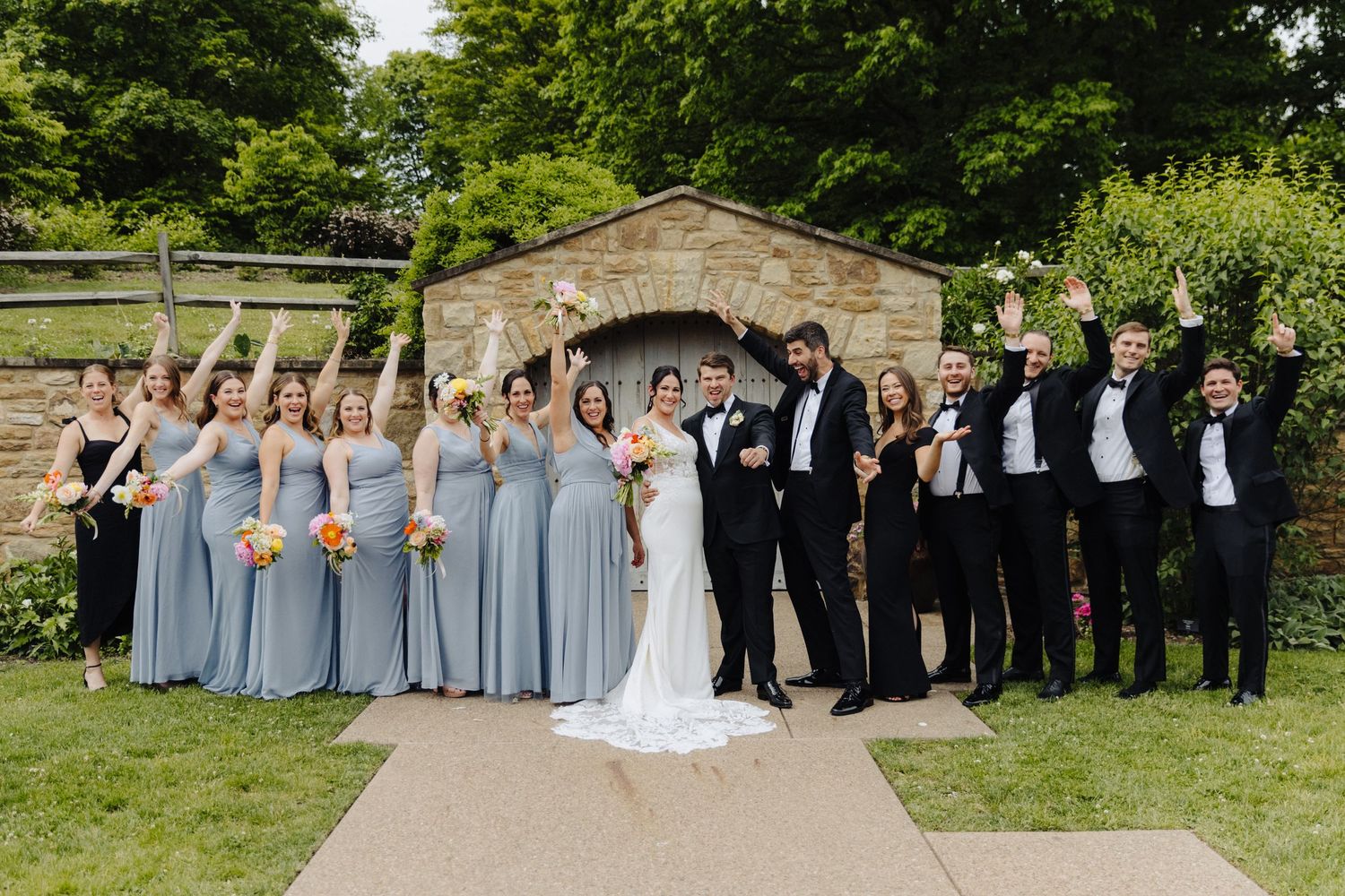 Wedding party in blue dresses and black tuxedos poses together on stone path.