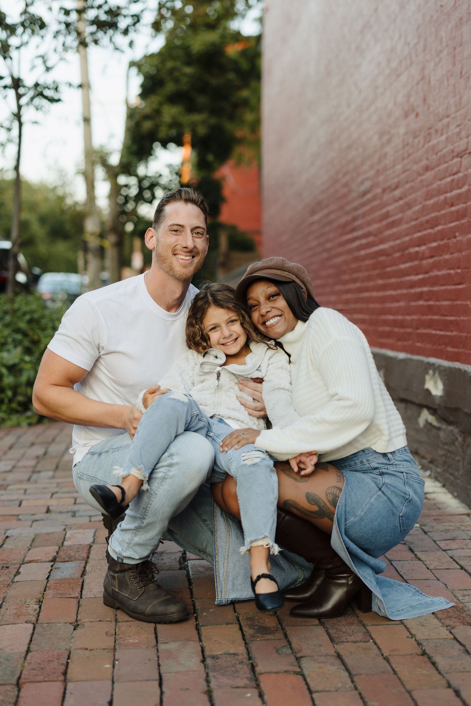 Couple in casual denim outfits sitting and laughing together on brick pavement by red wall.
