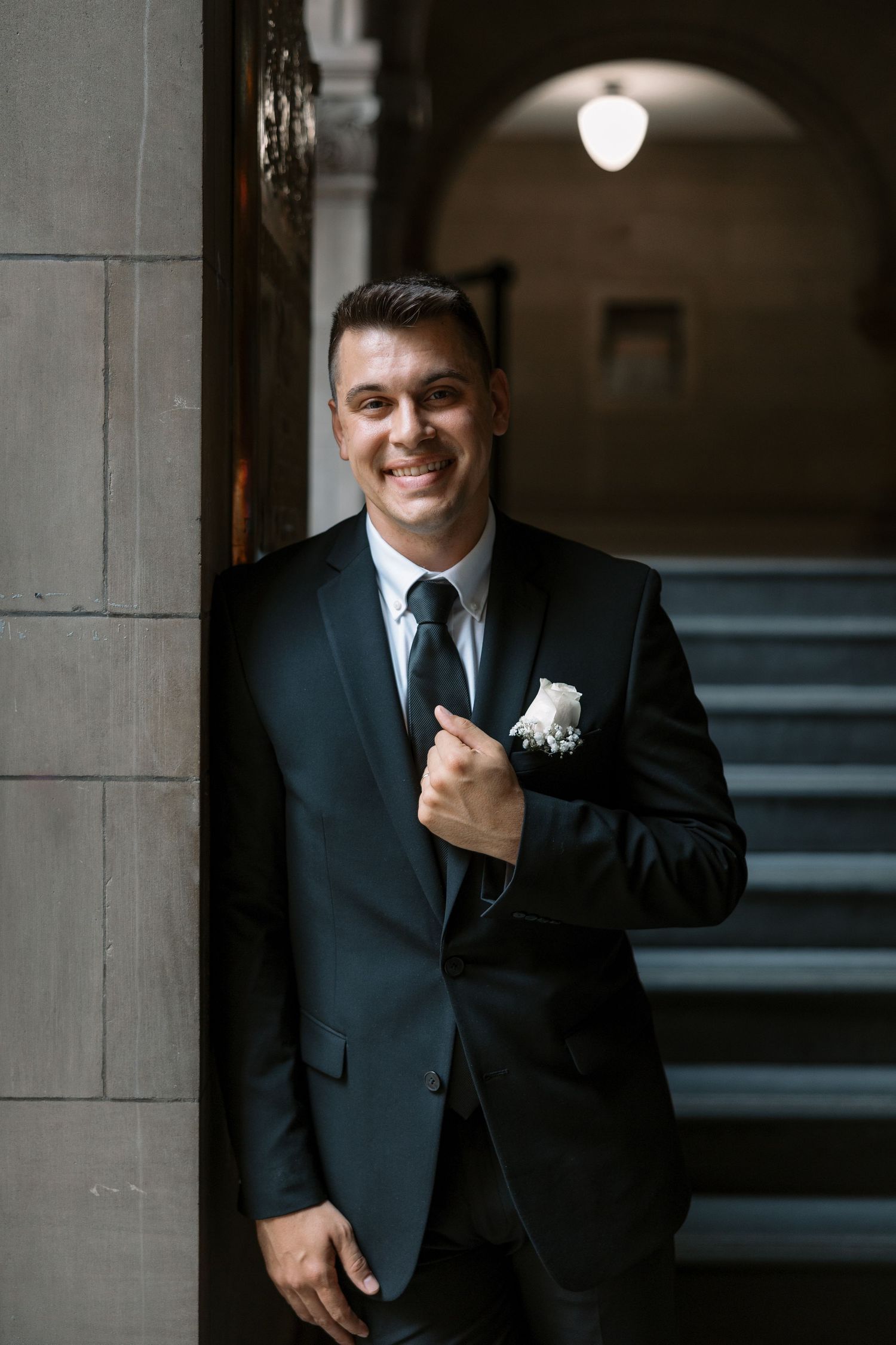 A groom in a dark suit poses against a stone column with classic architecture.