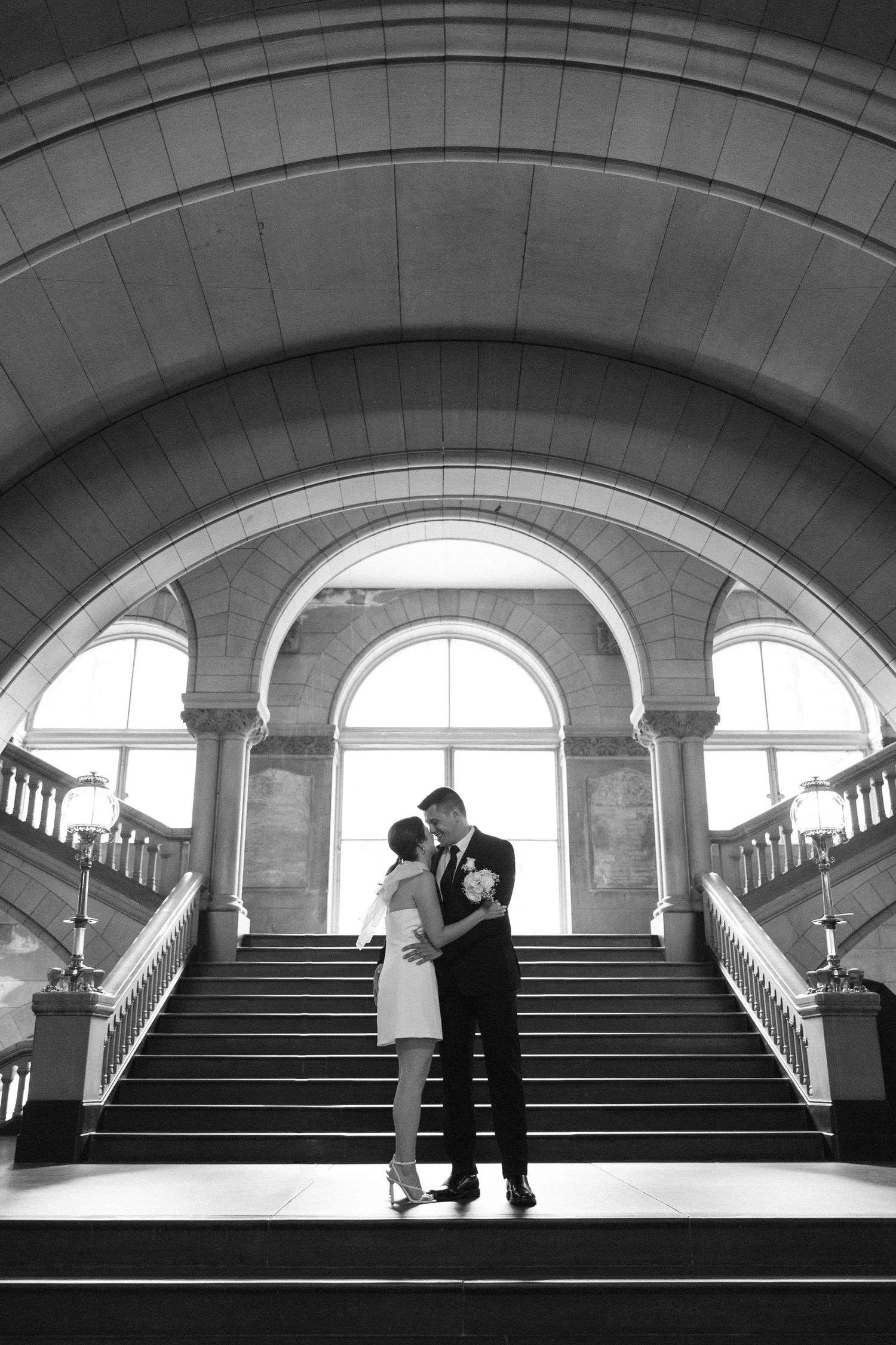 Couple shares intimate moment on grand staircase beneath arched windows in black and white.