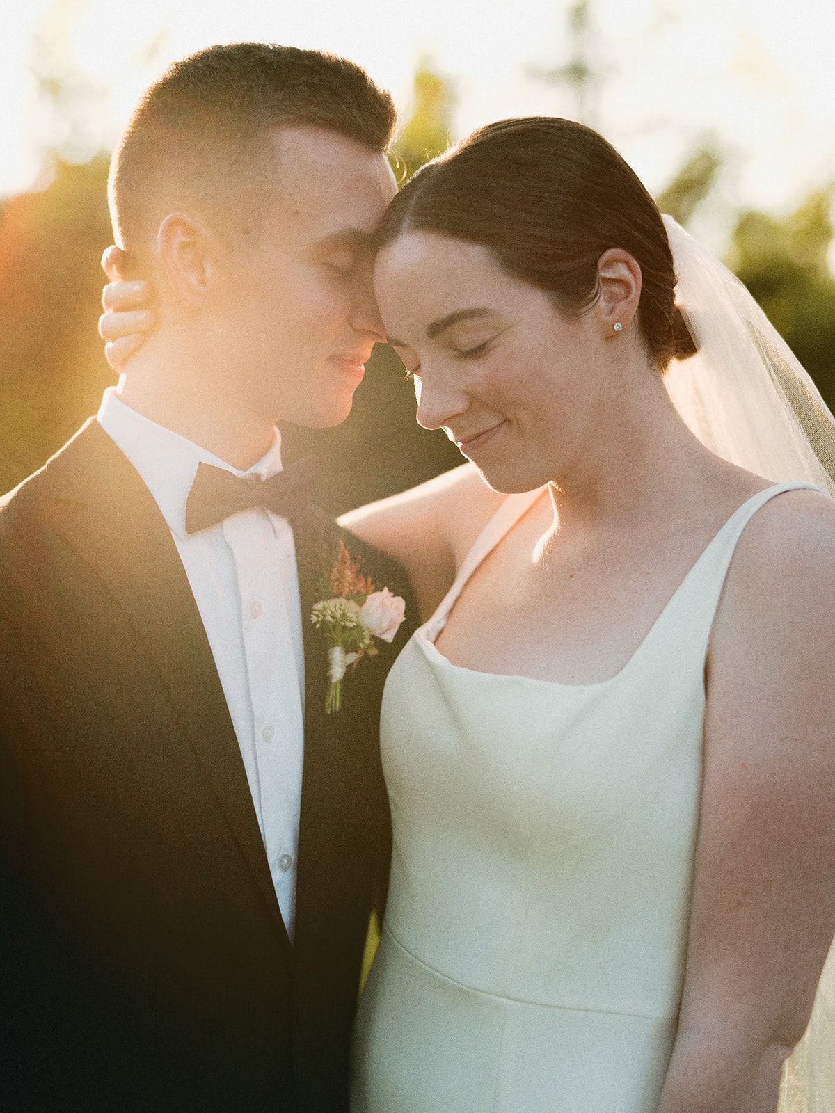 Golden hour wedding portrait of bride in white gown and groom in black tuxedo touching foreheads romantically outdoors.