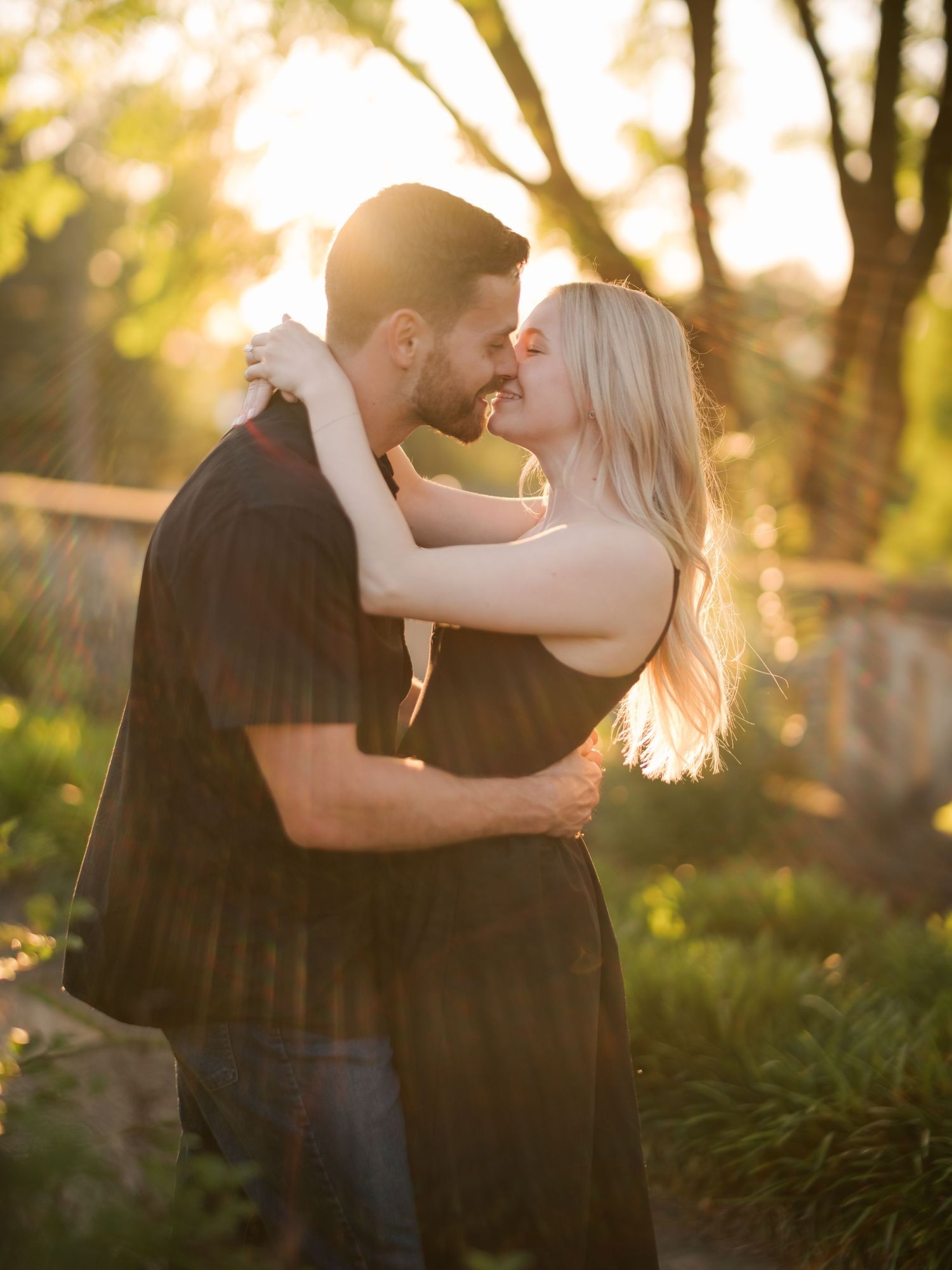 Romantic couple embraces and kisses in a sunlit garden at golden hour with soft bokeh through trees.