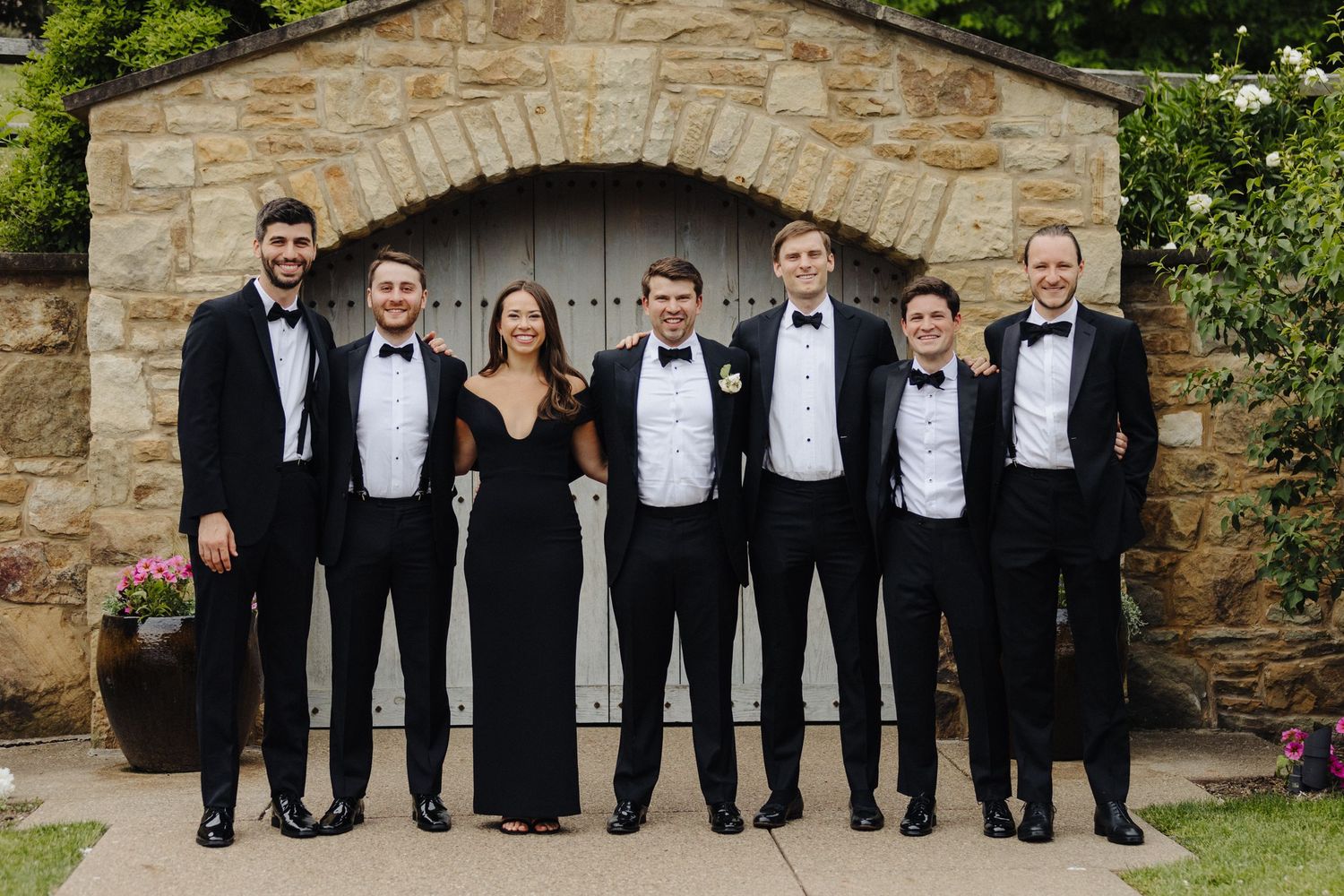 Wedding party lined up in formal black tuxedos and one black dress against stone archway.