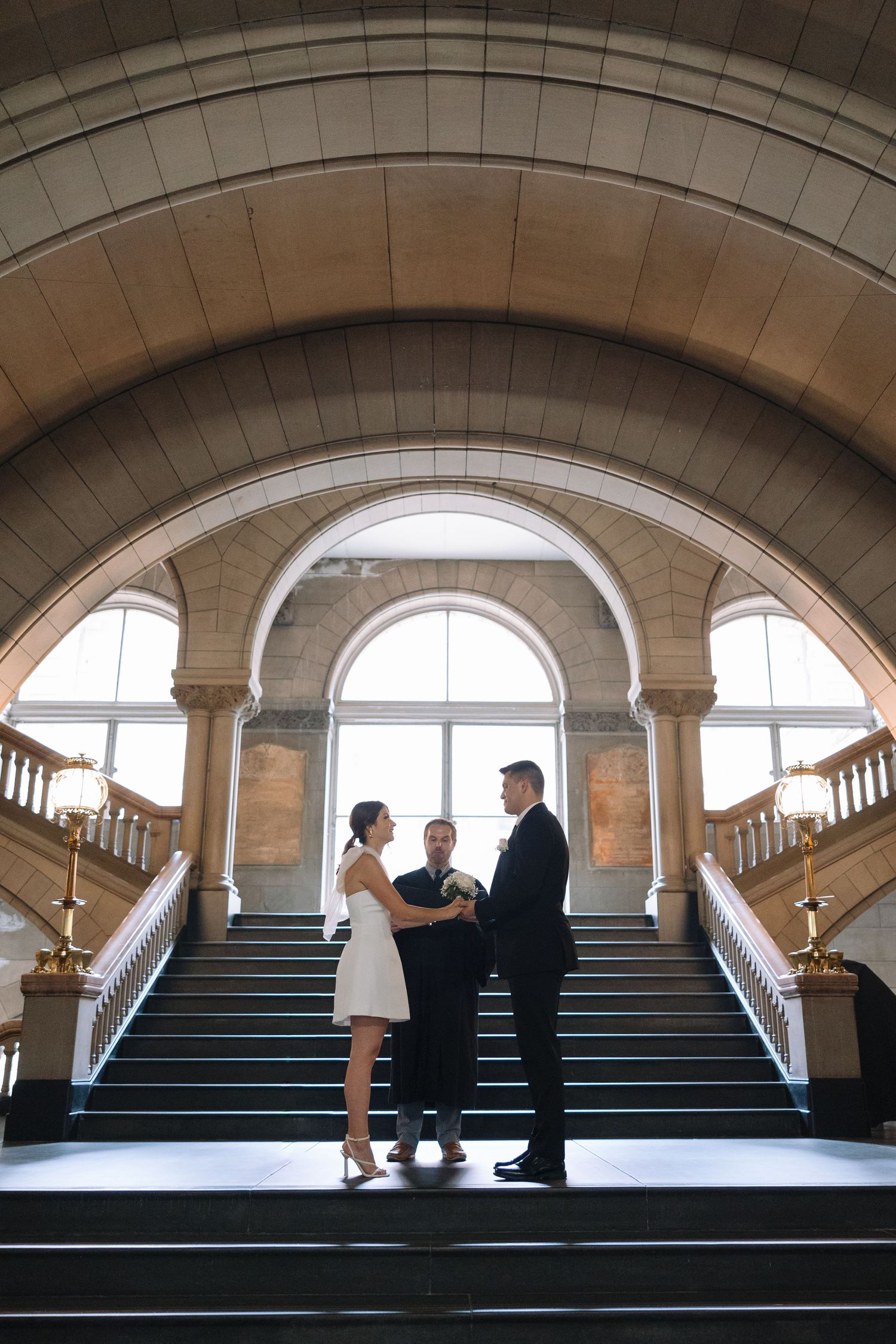 A wedding ceremony captured from above on grand marble stairs with dramatic arched windows.