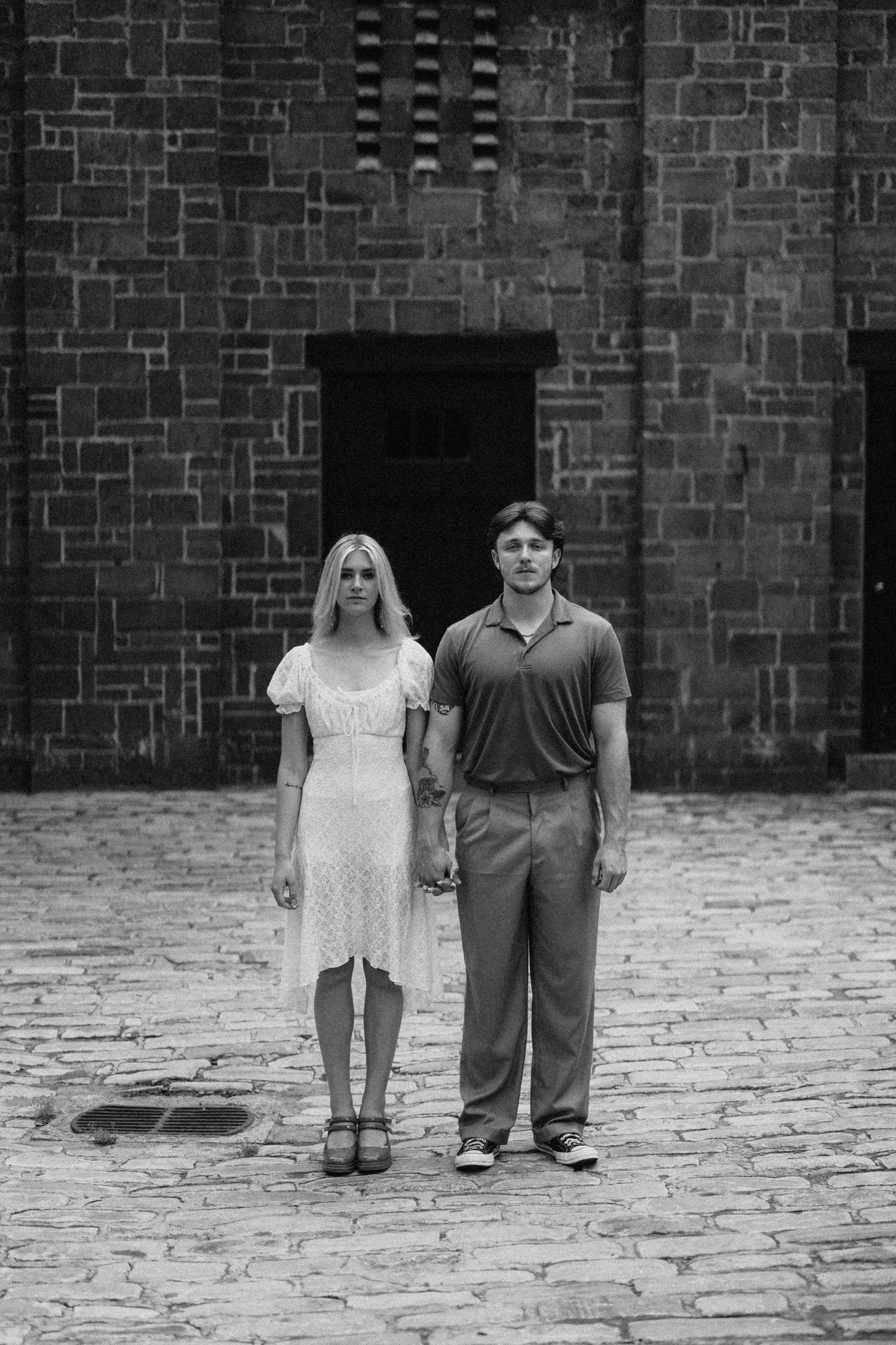 A black and white portrait of a couple standing on cobblestone against a brick wall.