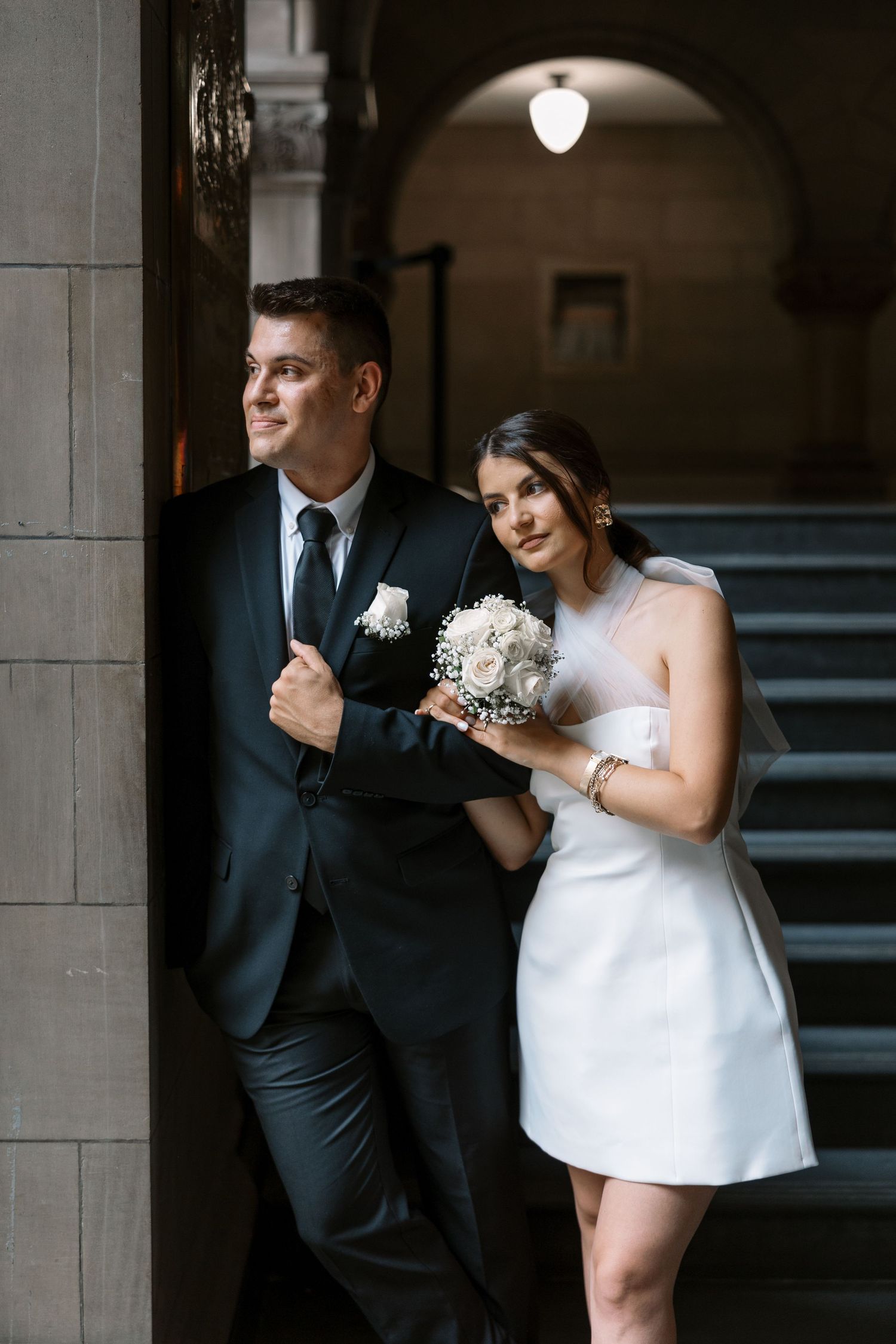 A couple poses elegantly against a stone column in a historic building with arched ceilings.