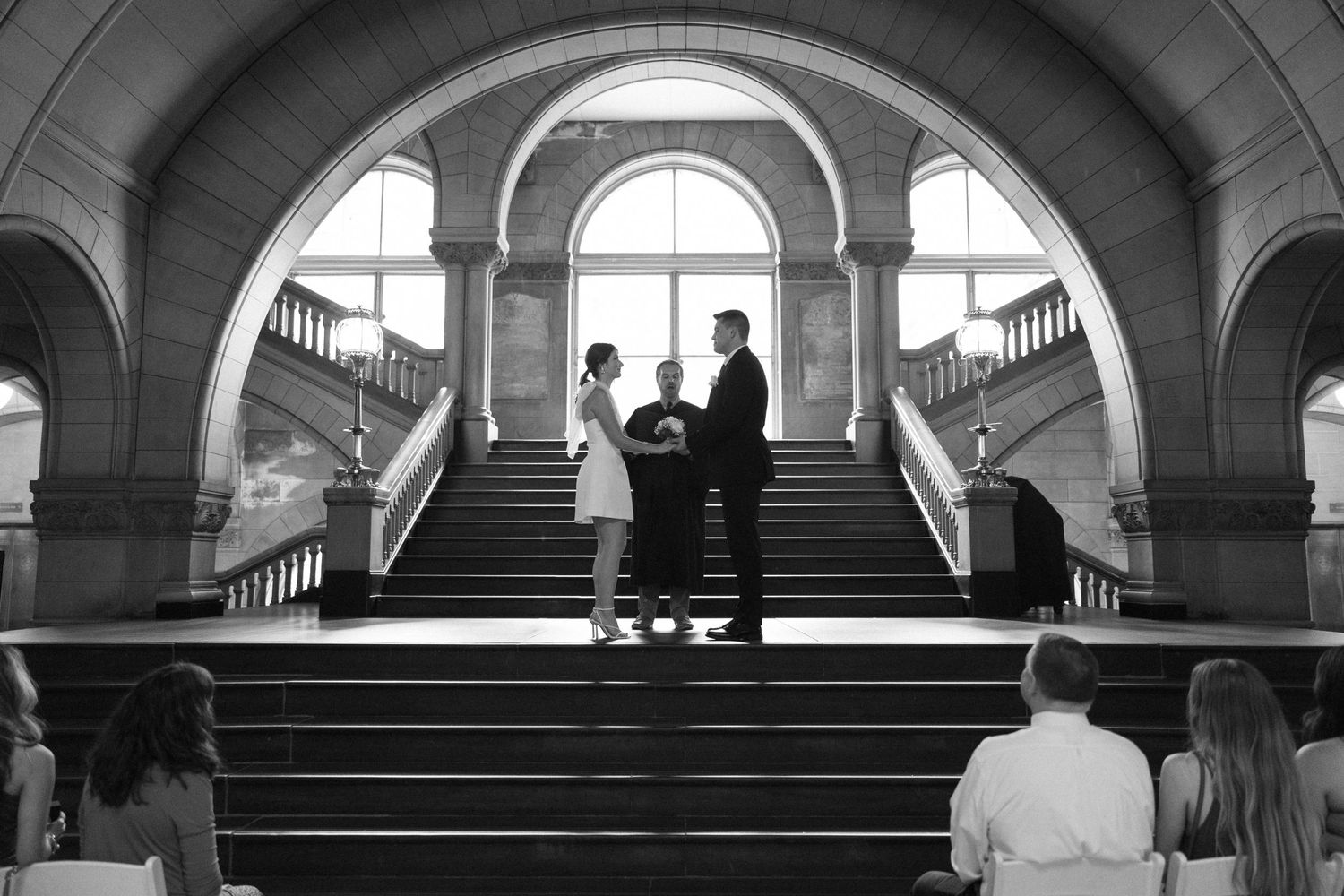 A black and white photograph of a ceremony taking place on grand marble stairs beneath an arched window.