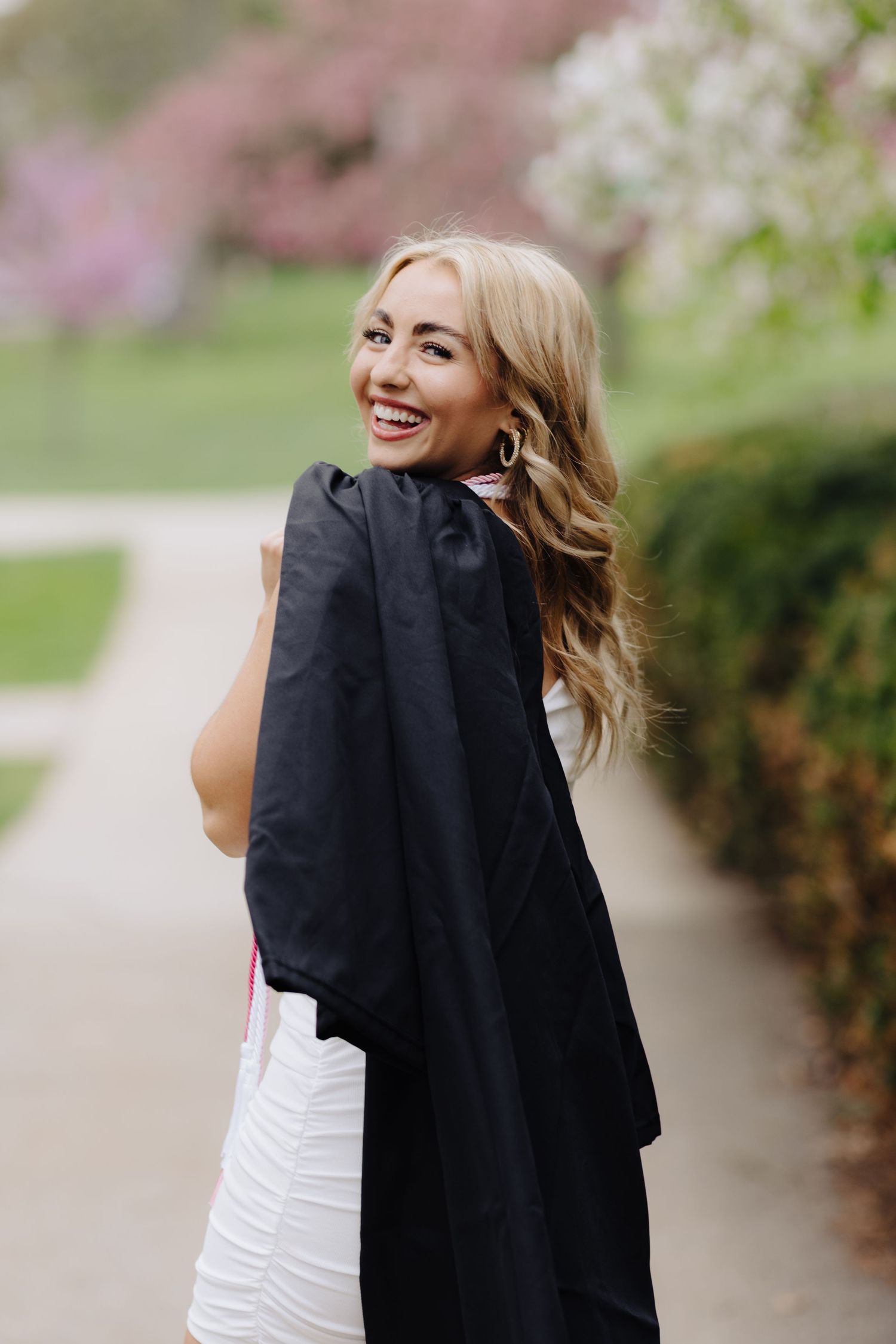 Graduate in white dress carries black gown while smiling near spring blossoms.