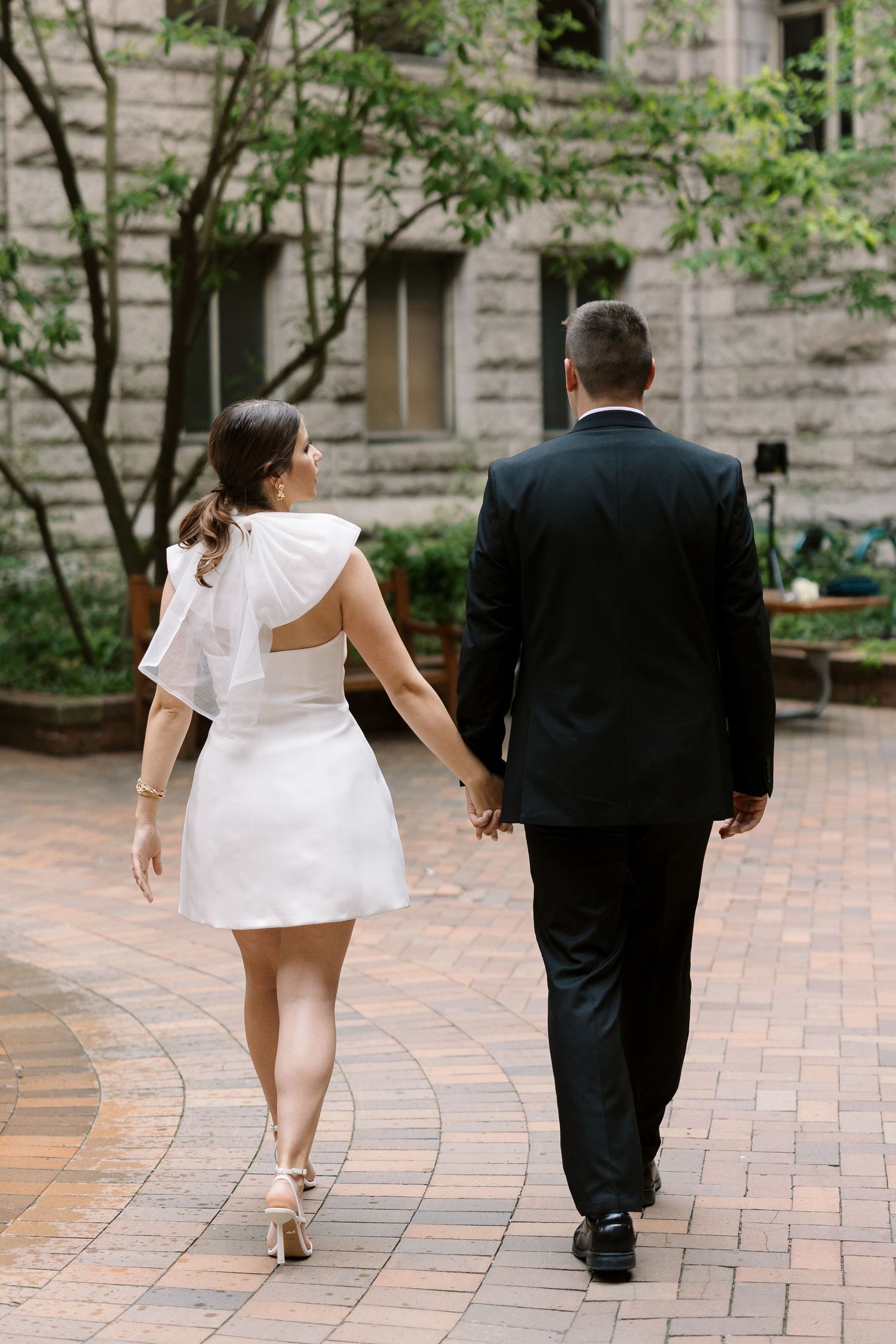A couple walks hand in hand along a brick pathway beside a stone building.
