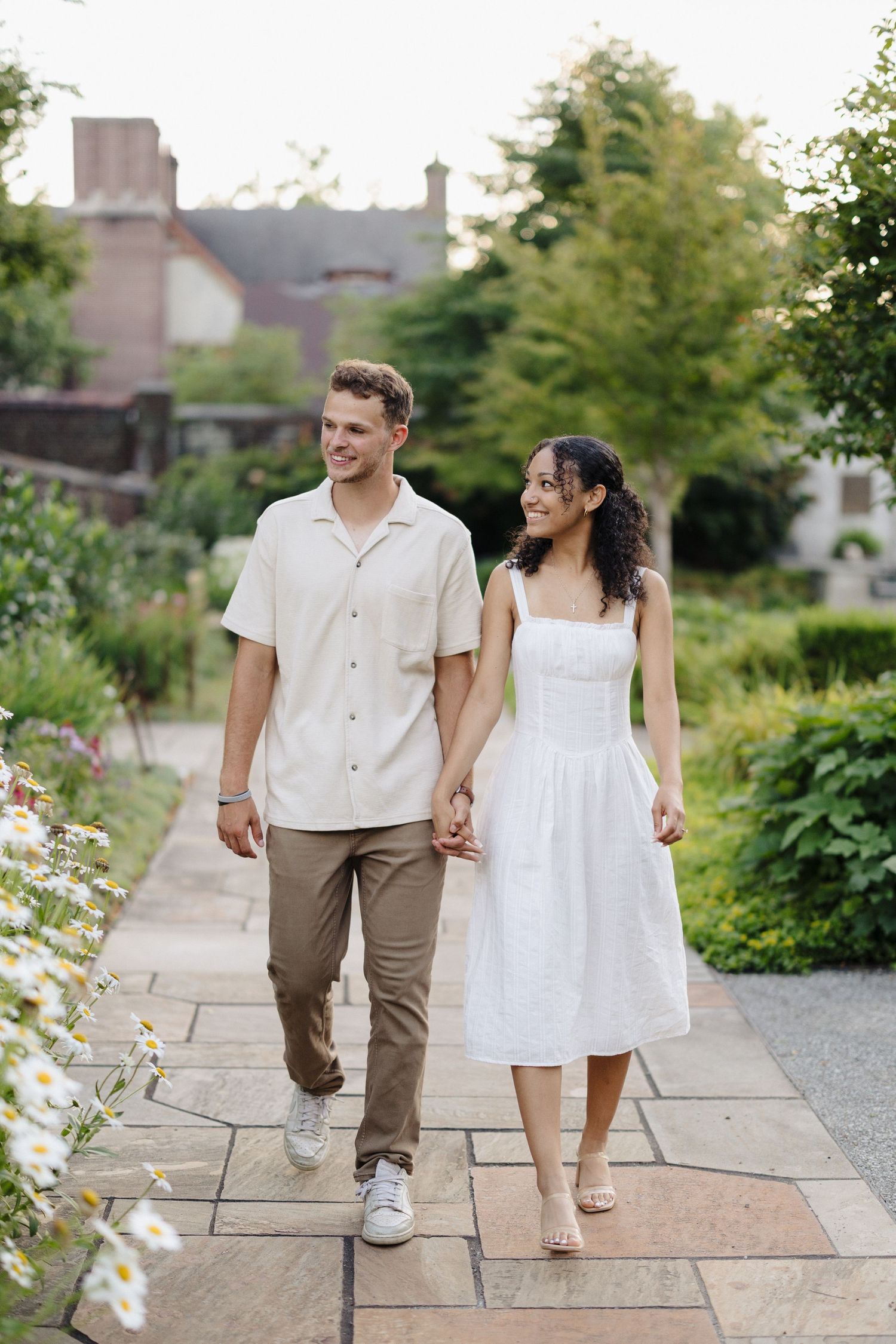 A couple walks hand in hand along a stone pathway surrounded by green landscaping and trees.