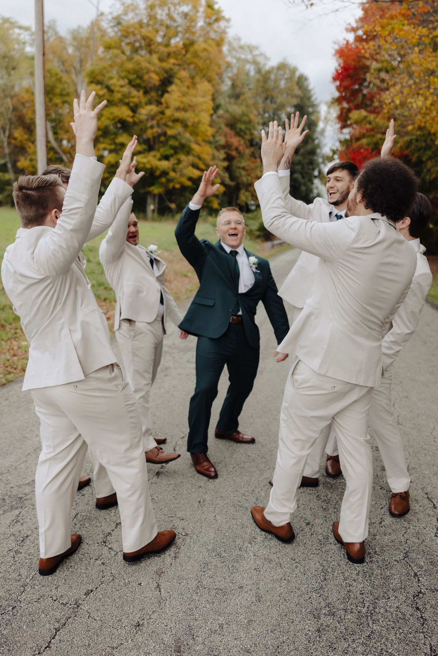 Groomsmen in light colored suits jump and celebrate with arms raised against autumn foliage background.