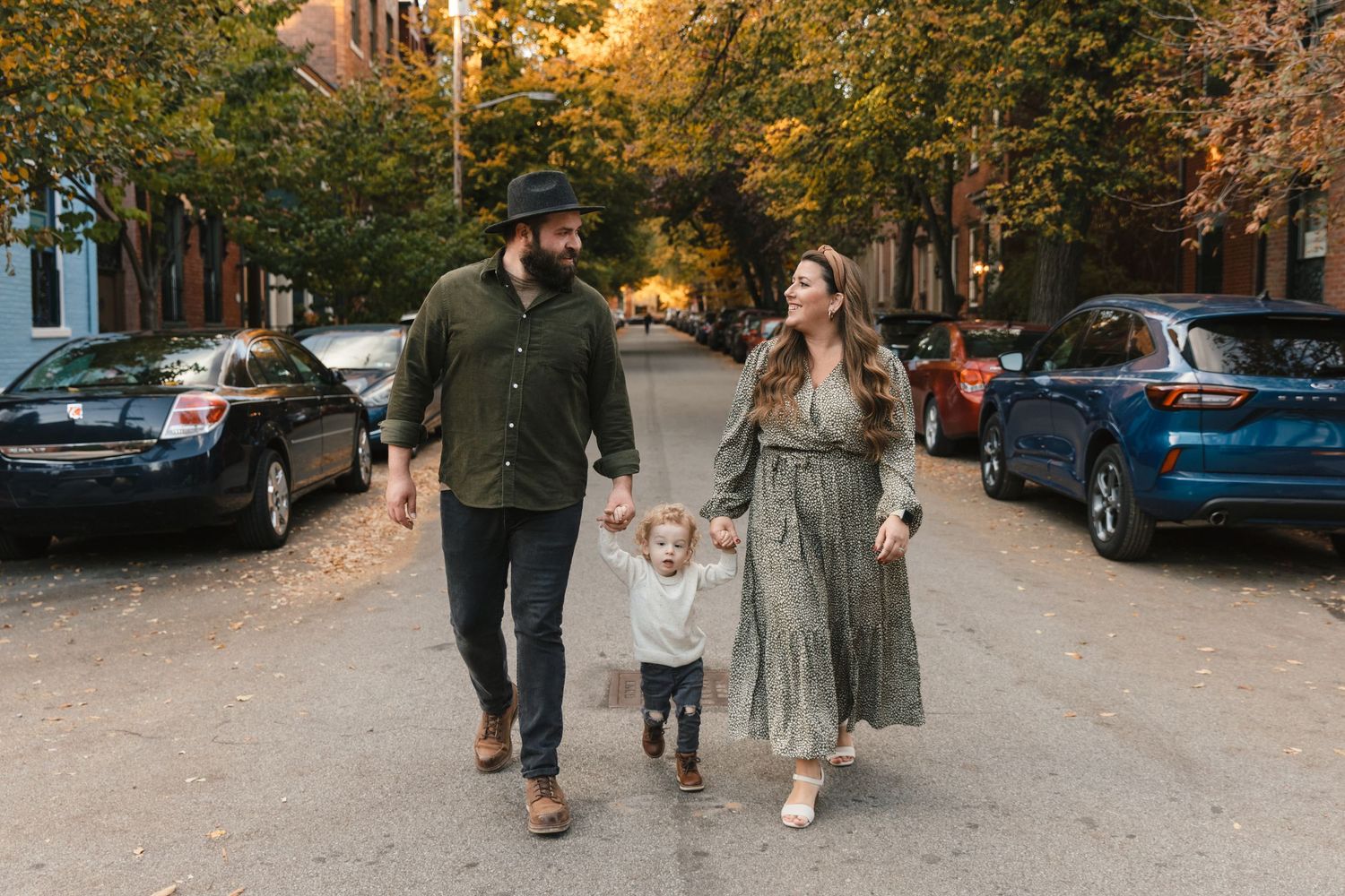 Family walking down tree-lined street lined with brownstone buildings in fall.