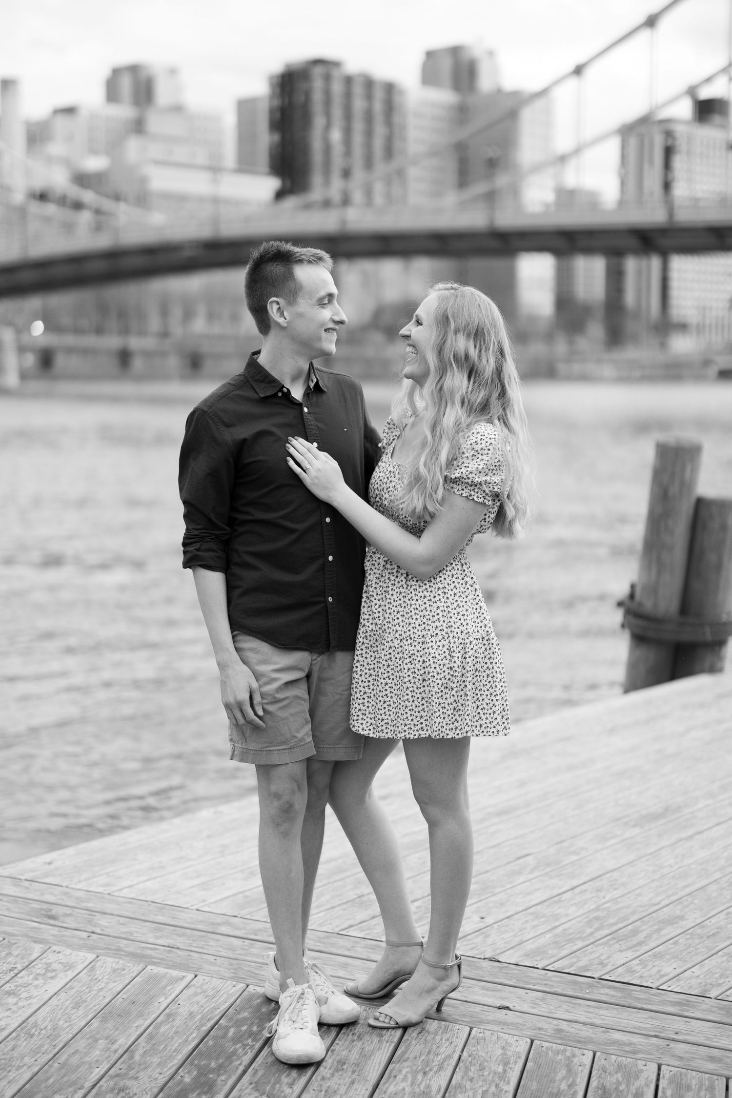 A black and white photo of a couple sharing a laugh on a wooden pier with a bridge behind them.