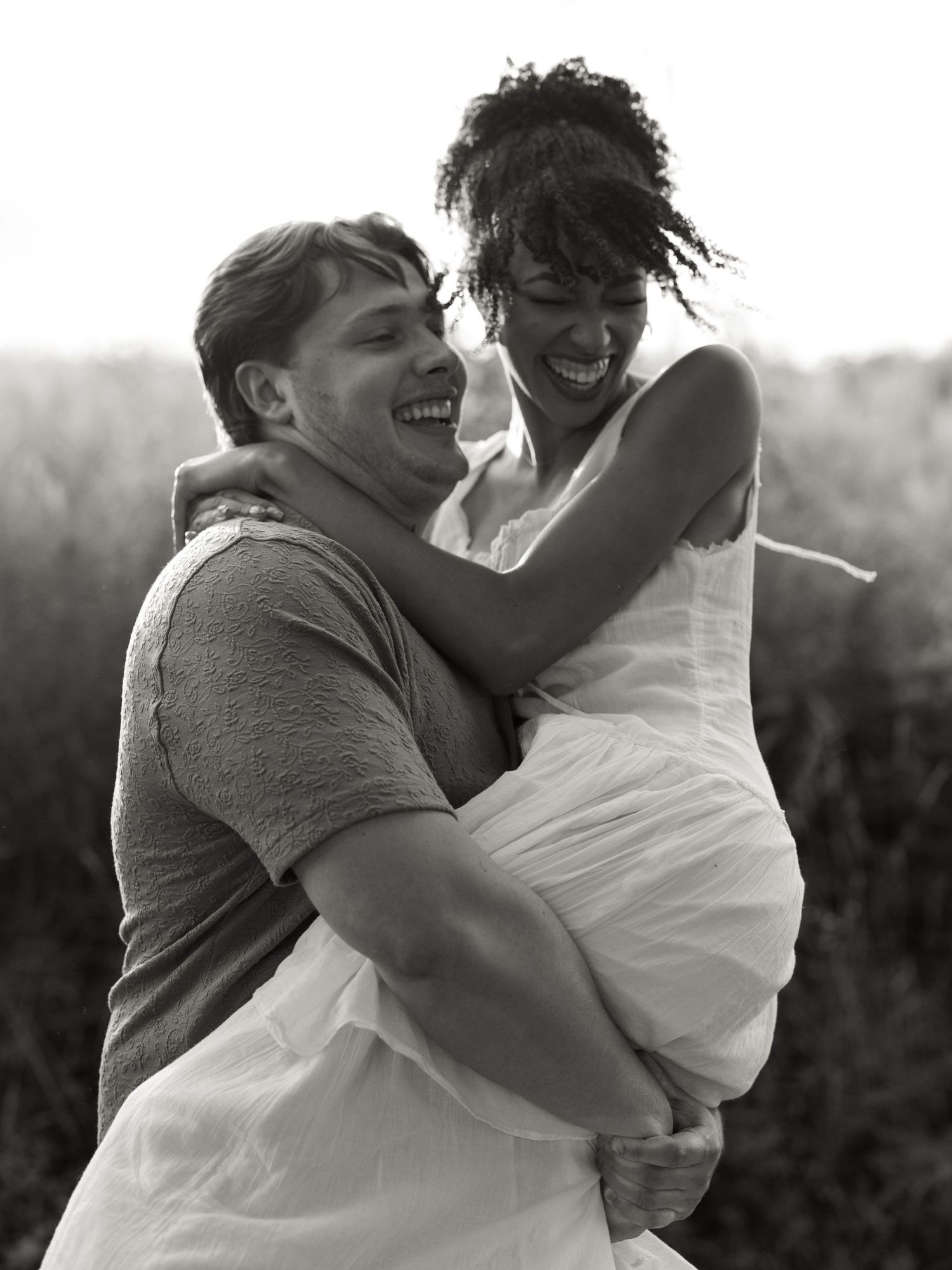 A black and white candid photograph captures a joyful couple embracing and laughing together outdoors.