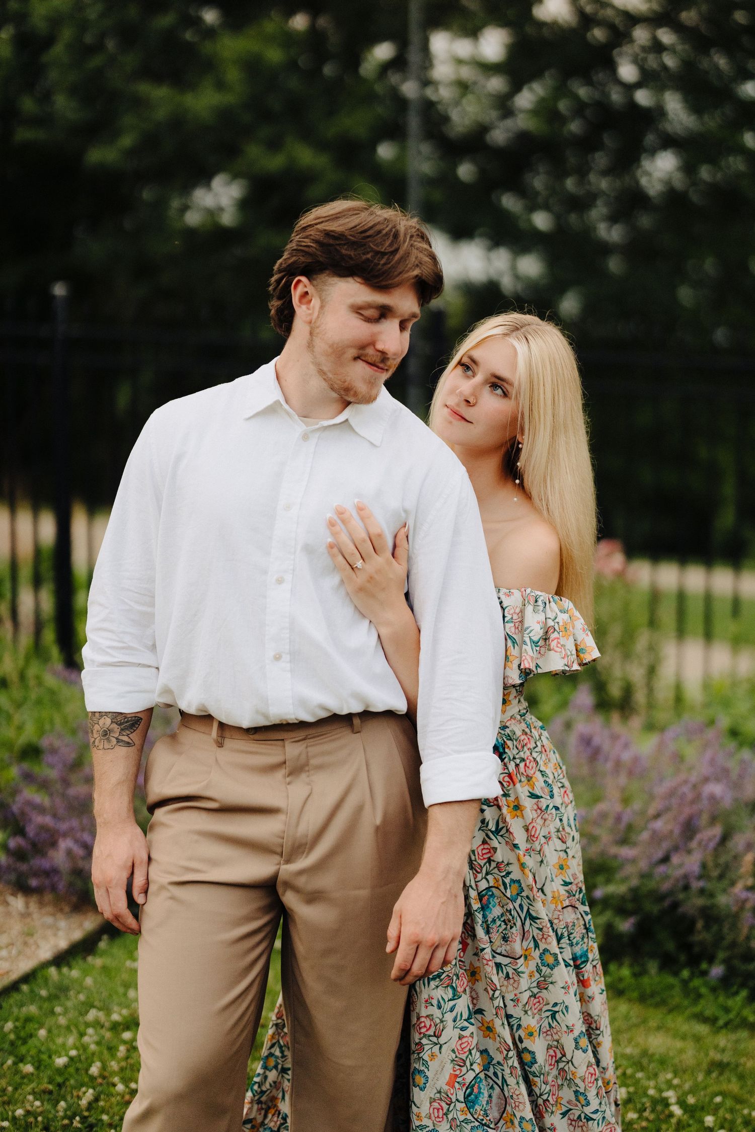 Couple poses intimately in garden with floral dress and classic white shirt.
