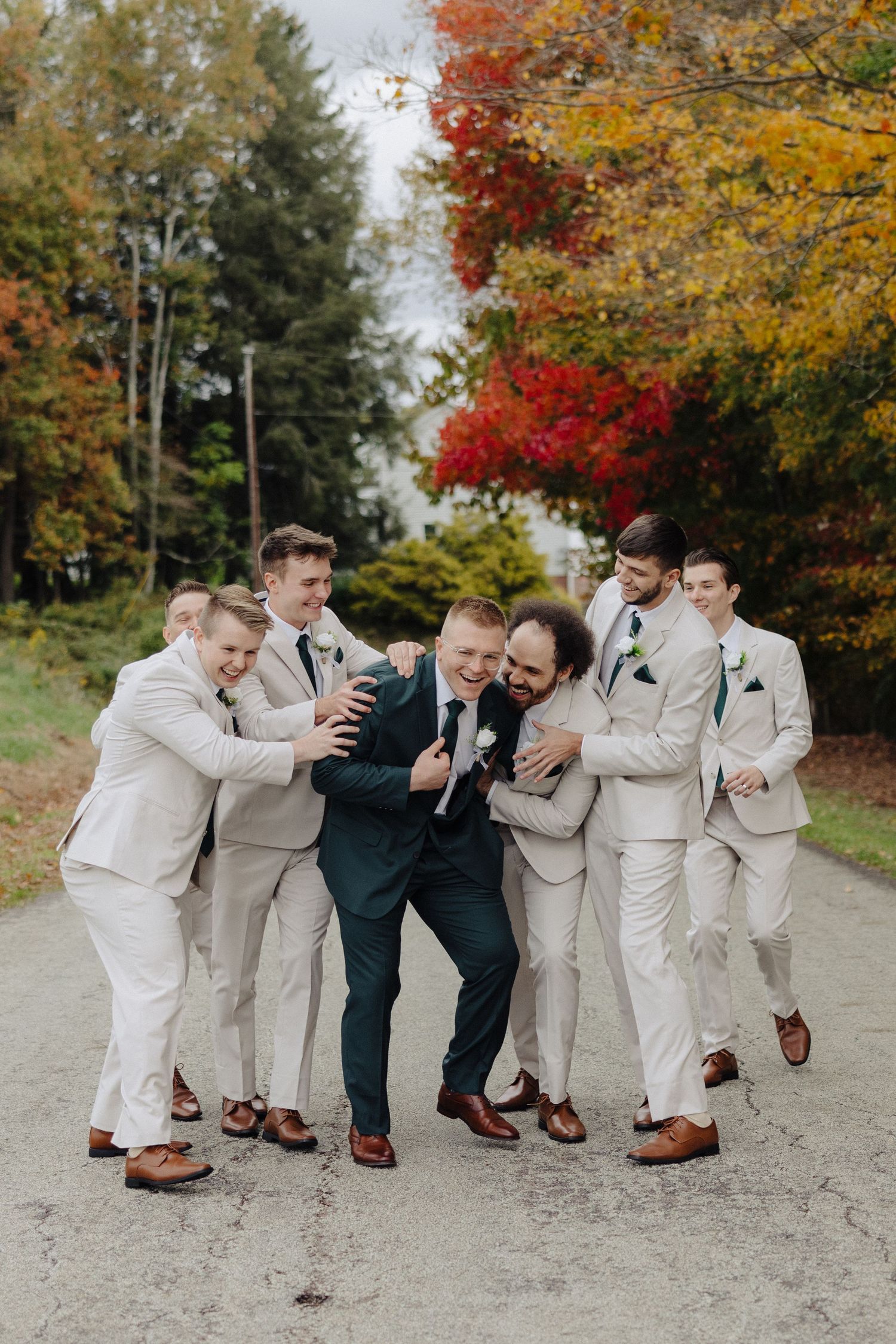 Groomsmen in light suits playfully pose with groom in navy suit against autumn trees.