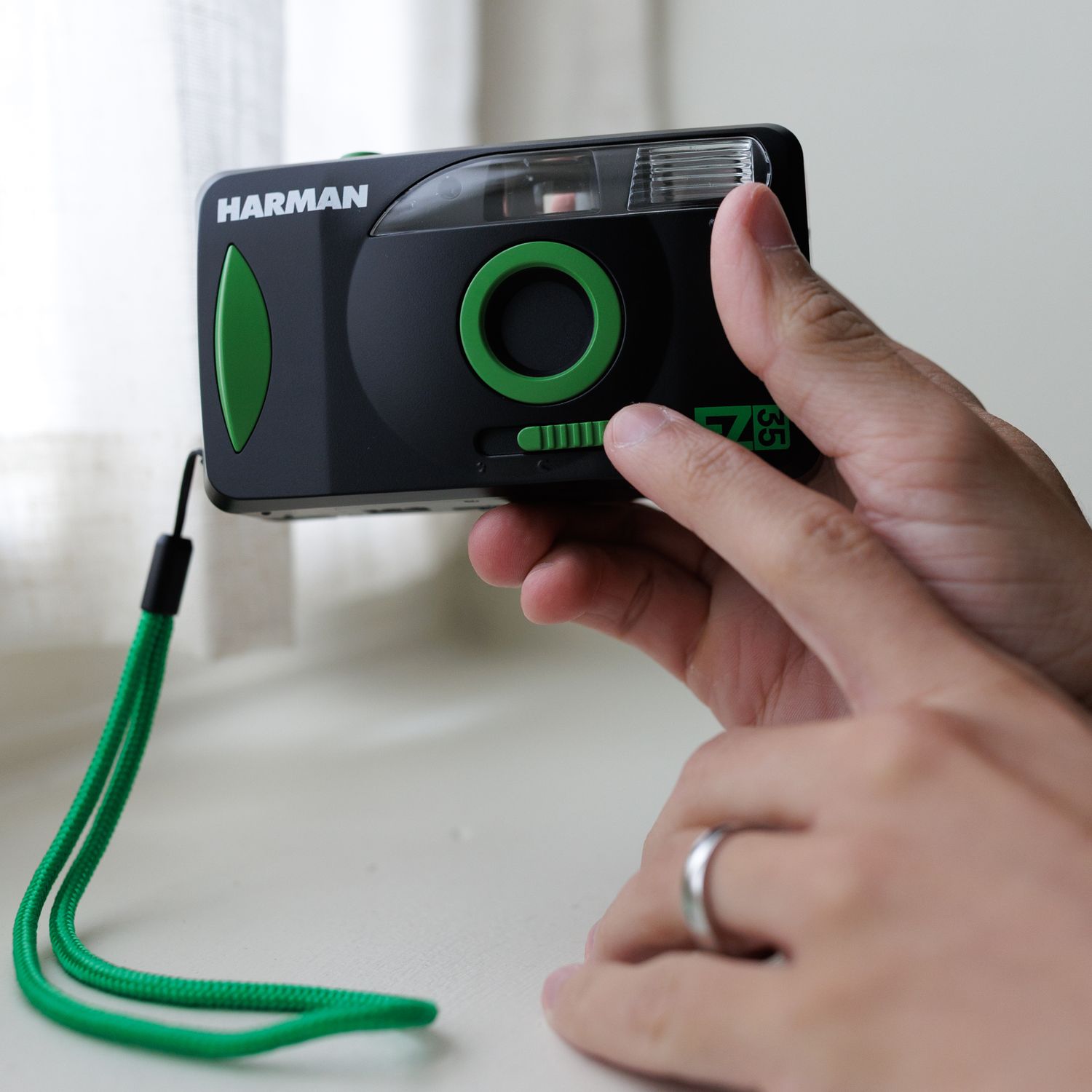 Black Harman point-and-shoot film camera with green accents and wrist strap held in someone's hand.