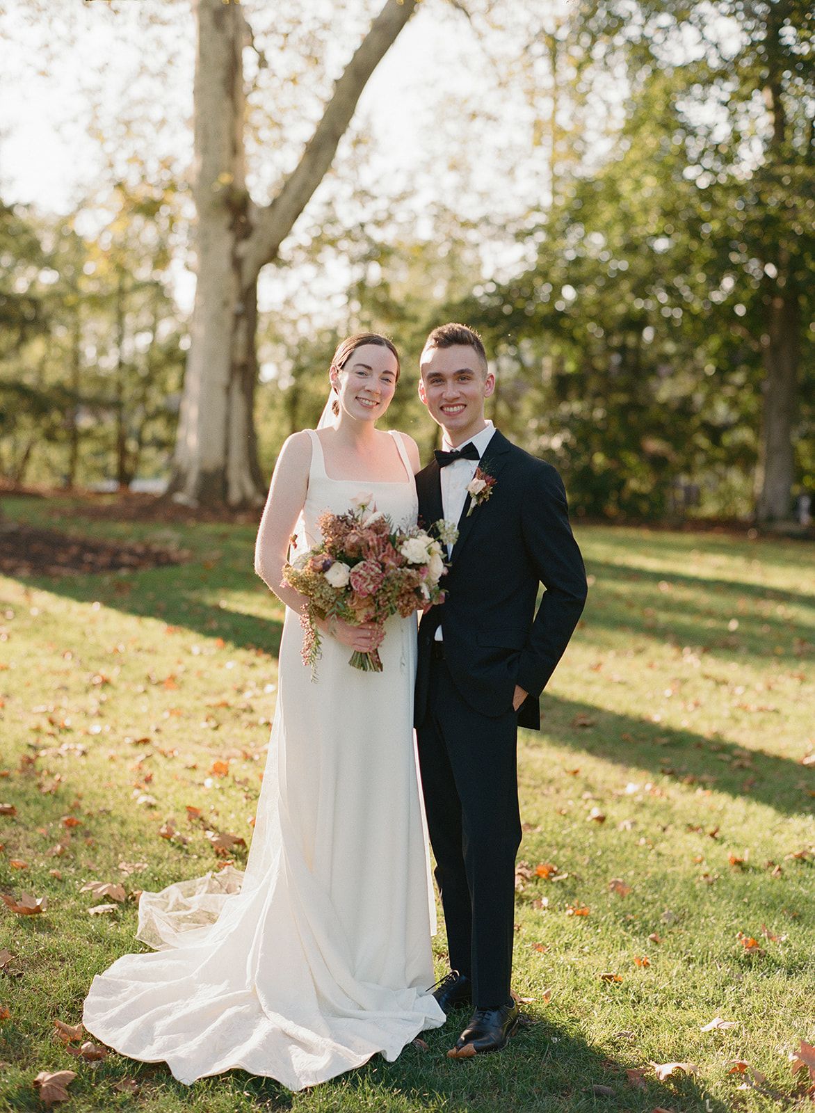 Bride in white gown holding floral bouquet stands with groom in black tuxedo on green lawn surrounded by autumn trees.