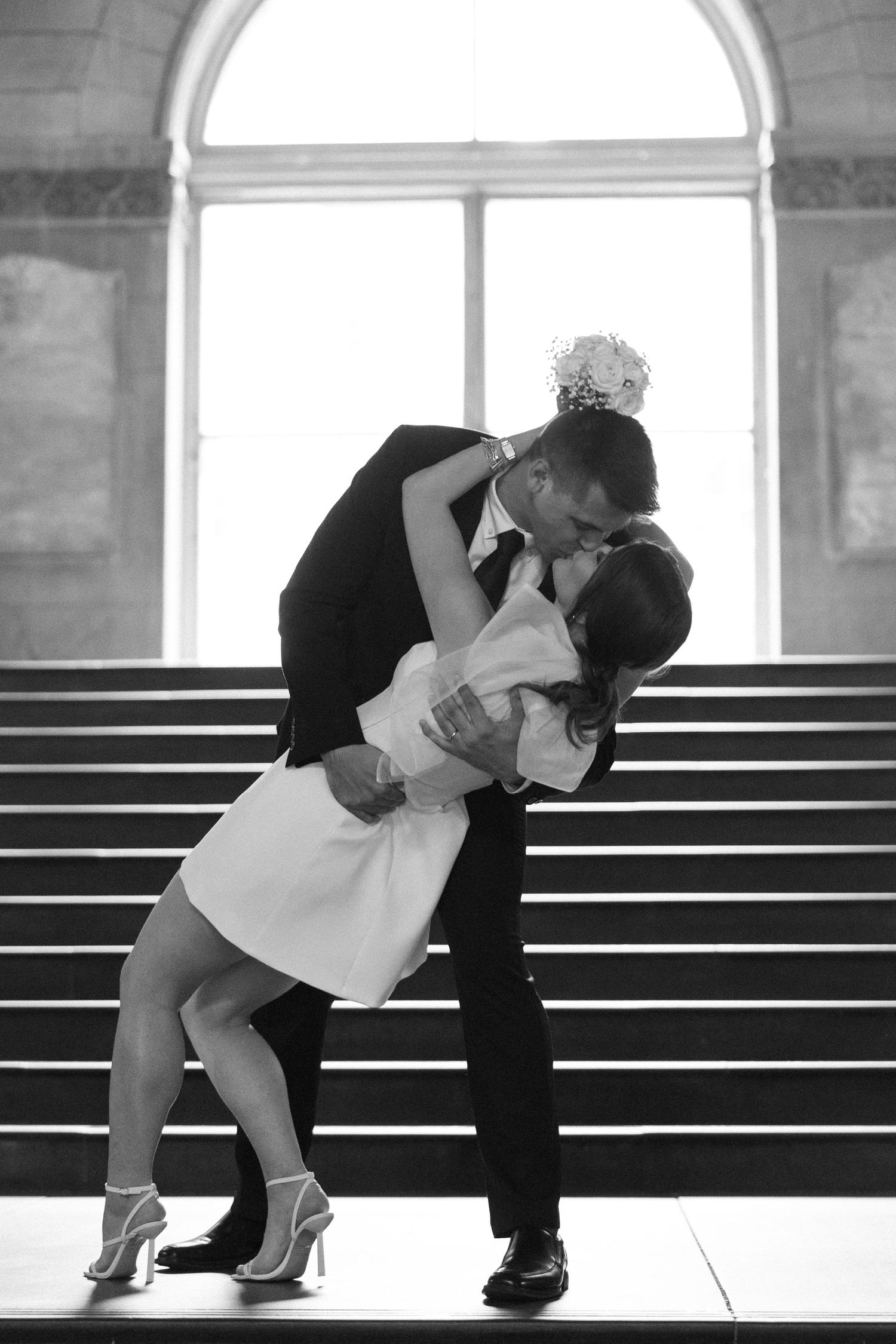 A romantic dip kiss on stone steps beneath an arched window in black and white.