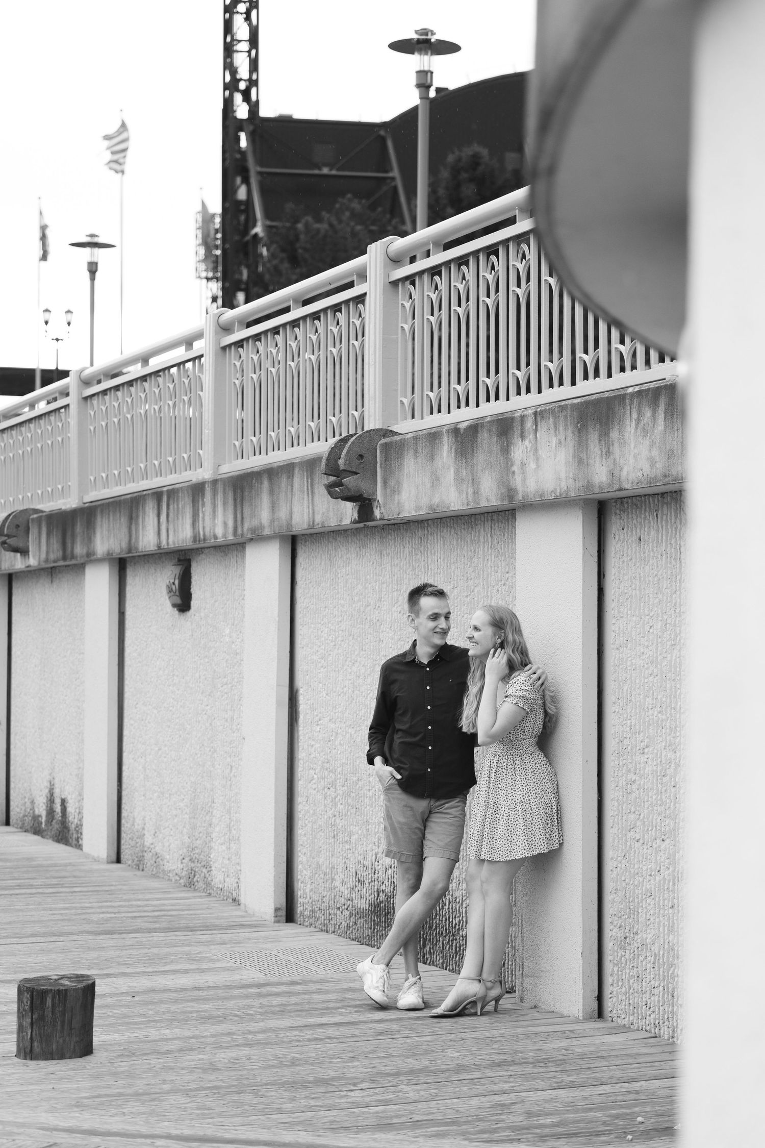 A black and white portrait of a couple leaning against a wall on a boardwalk.