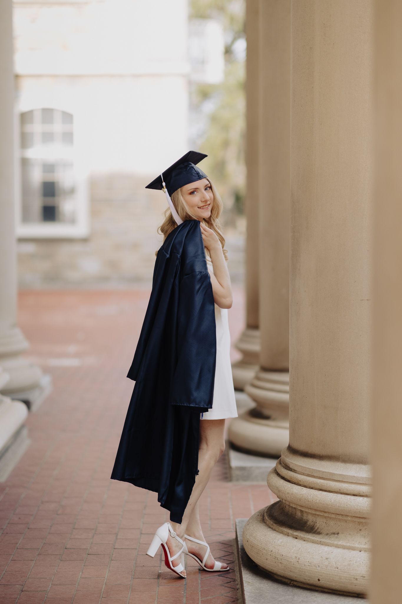 Graduate in cap and gown poses elegantly between classical stone columns.