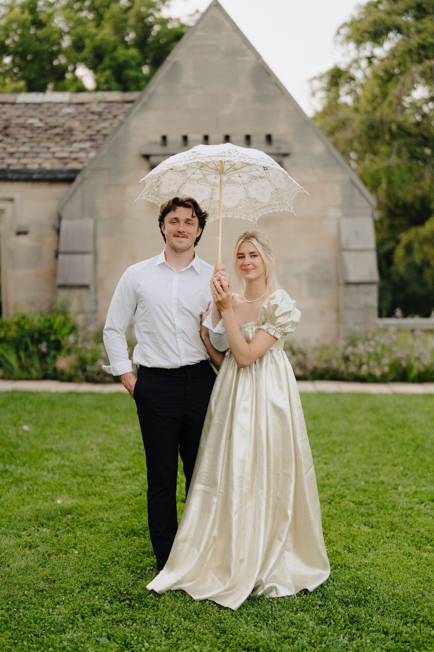 A couple shares a romantic moment under a white lace umbrella in front of a stone church on their wedding day.