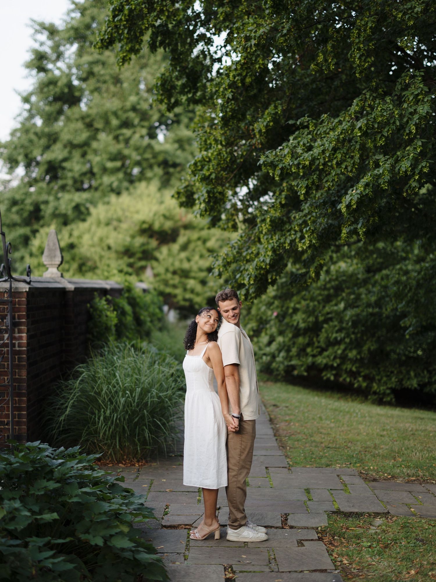 A couple in wedding attire walks together on a stone path surrounded by lush green trees and foliage.