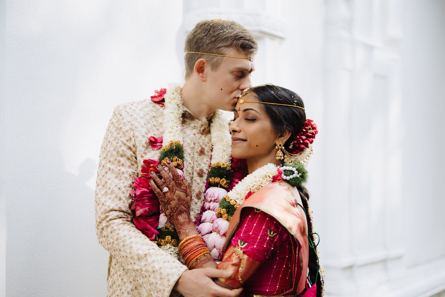 Wedding couple in traditional fusion attire adorned with red and pink floral garlands share an intimate moment.